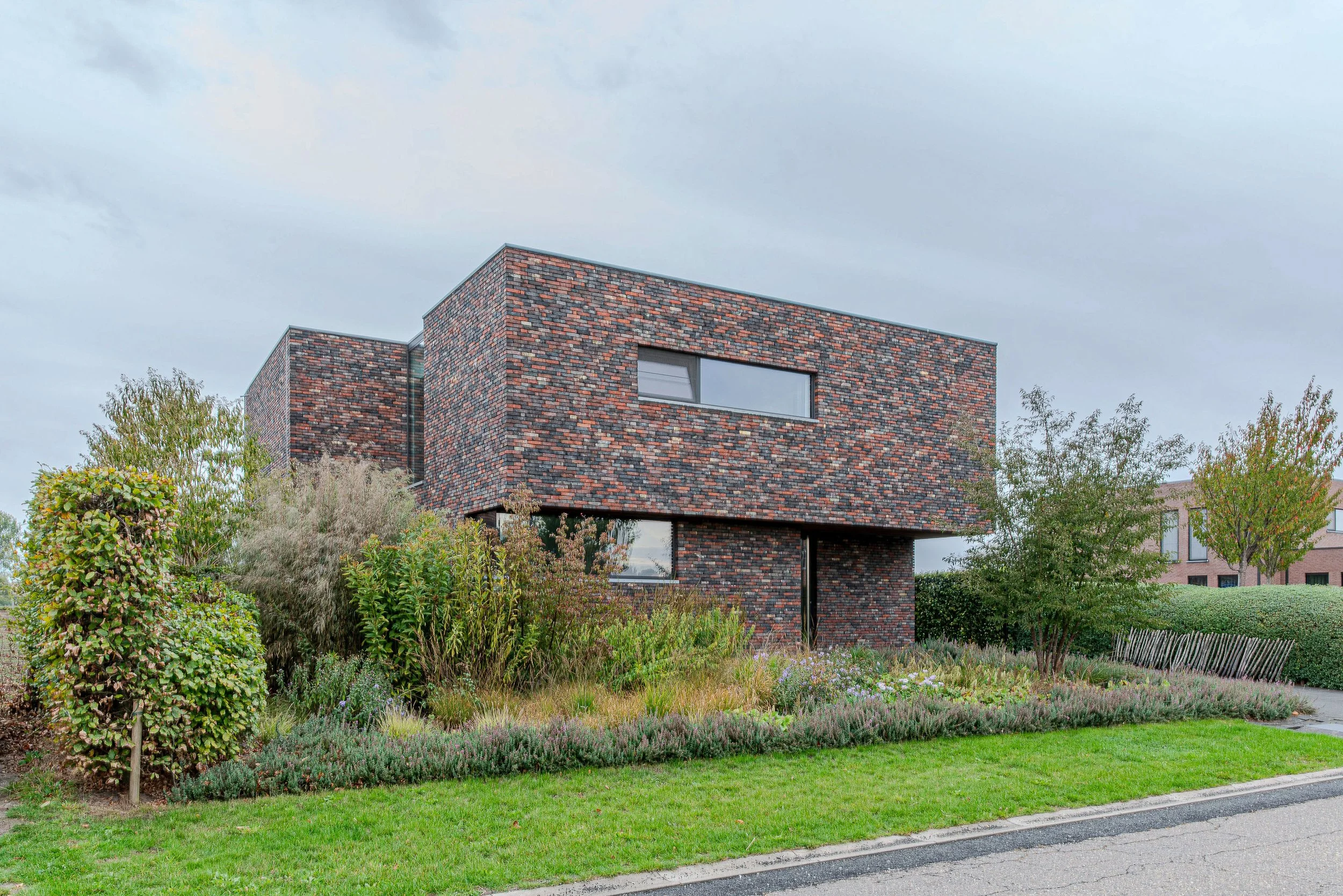 Modern huis met donkere bakstenen gevel en grote raam, omgeven door groene begroeiing en tuin ontworpen door Gert Anthonissen Architect.