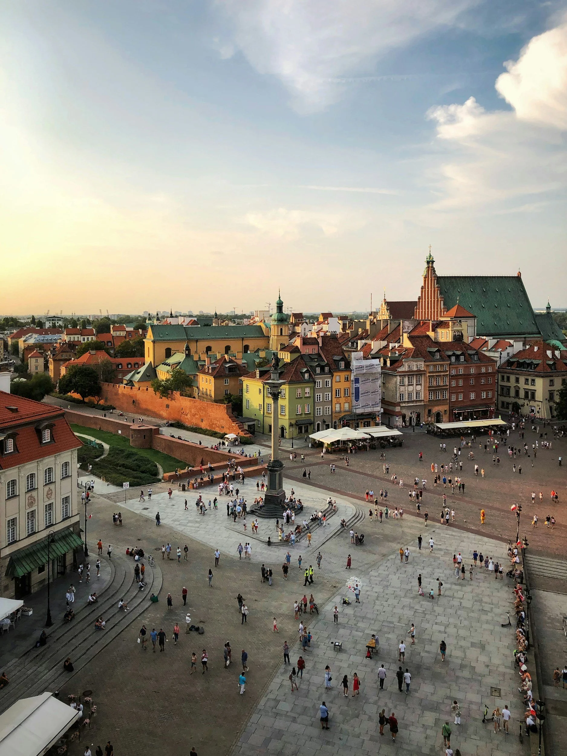 View of a city square in the evening with many people walking, historic buildings, and a statue in the center. The sky is partly cloudy and the sunlight is casting a warm glow.