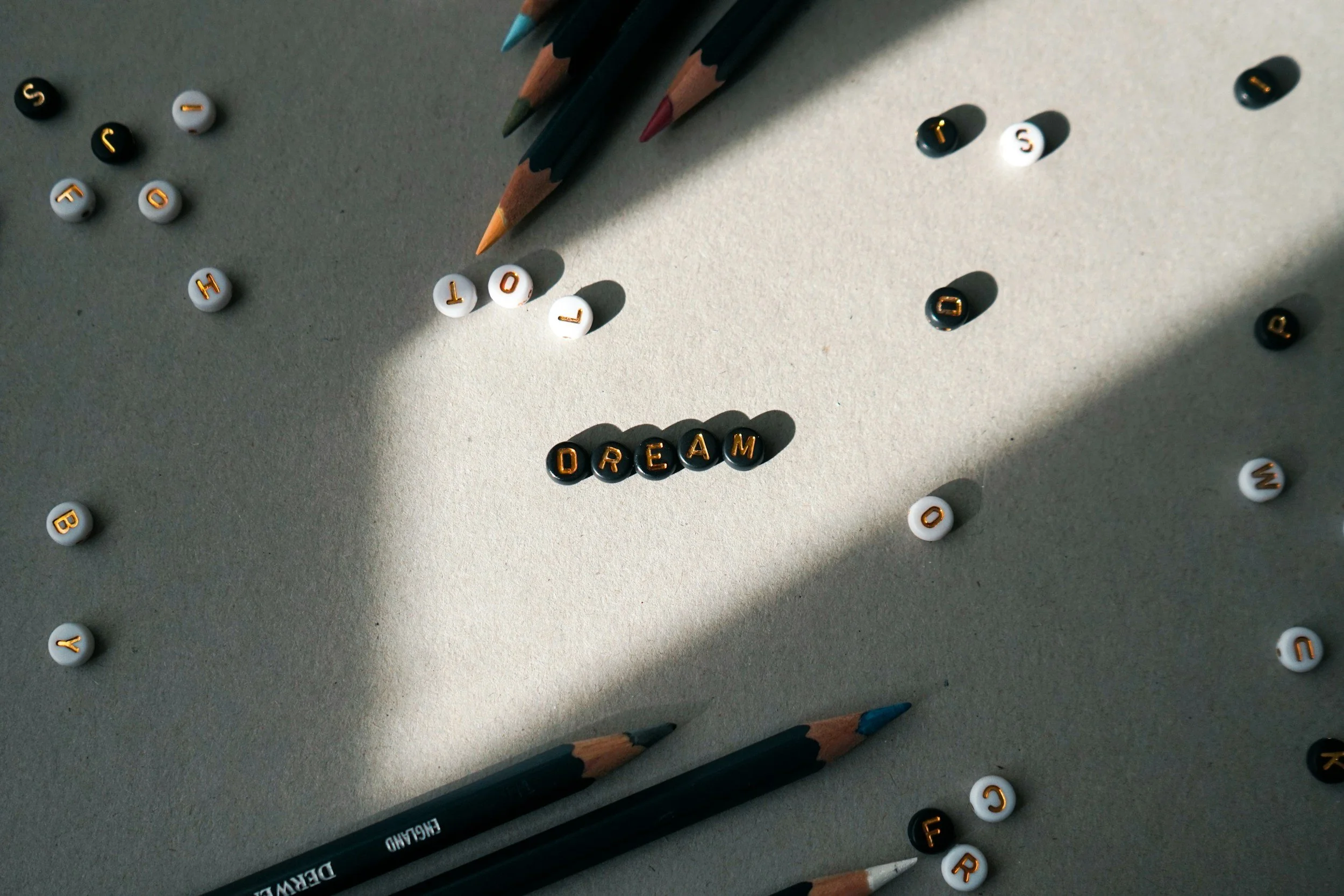 Colored pencils and scattered letter beads spelling the word 'DREAM' on a light gray surface, with sunlight casting shadows.