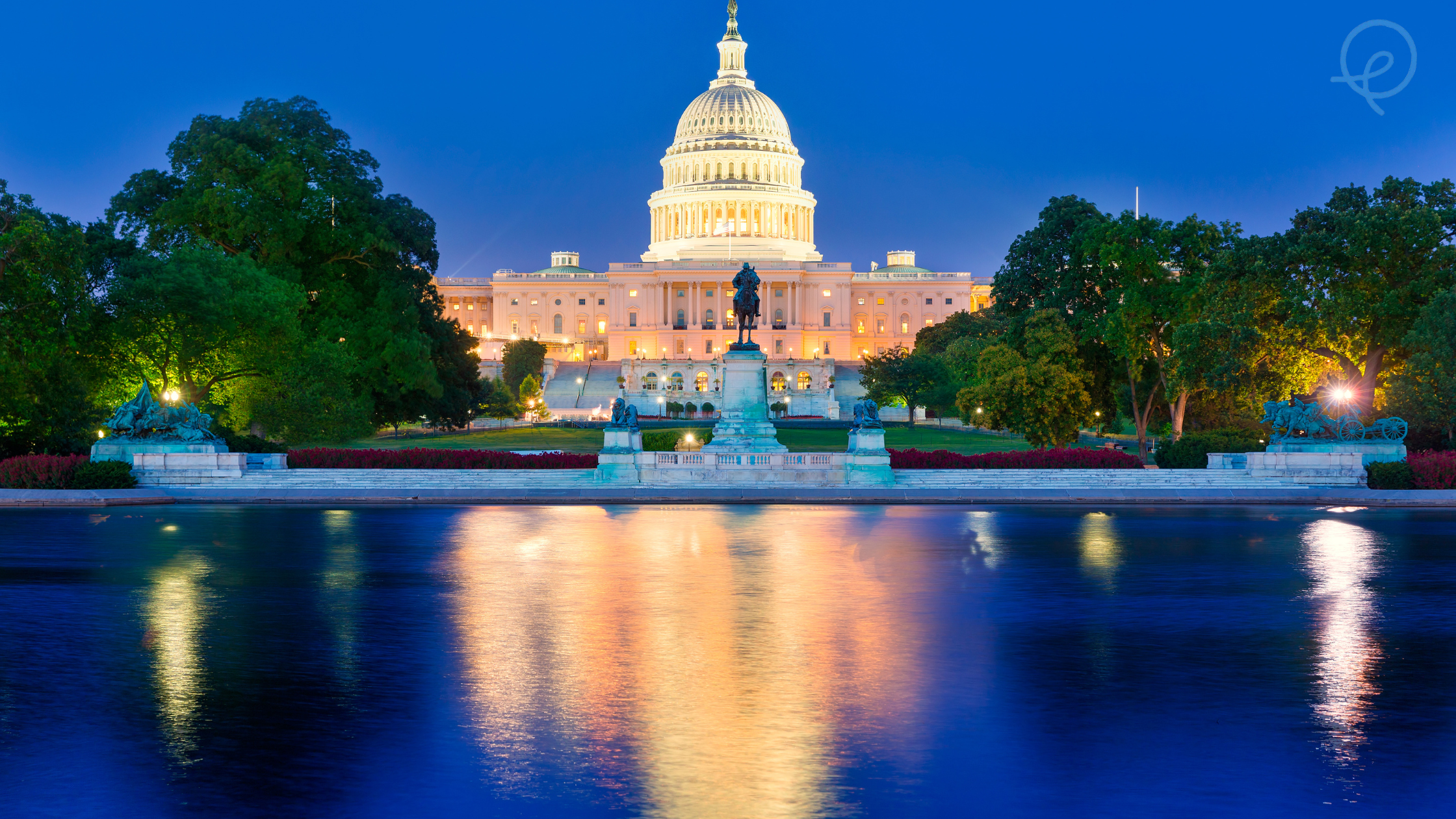 Night view of the U.S. Capitol building in Washington, D.C., illuminated and reflected in the water in front, with trees and sculptures in the foreground.