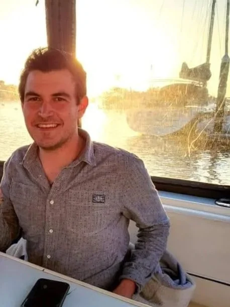 A smiling man with short dark hair, wearing a gray button-up shirt, sitting at a table on a boat or near water, with the sun setting in the background and a sailboat visible through a window.