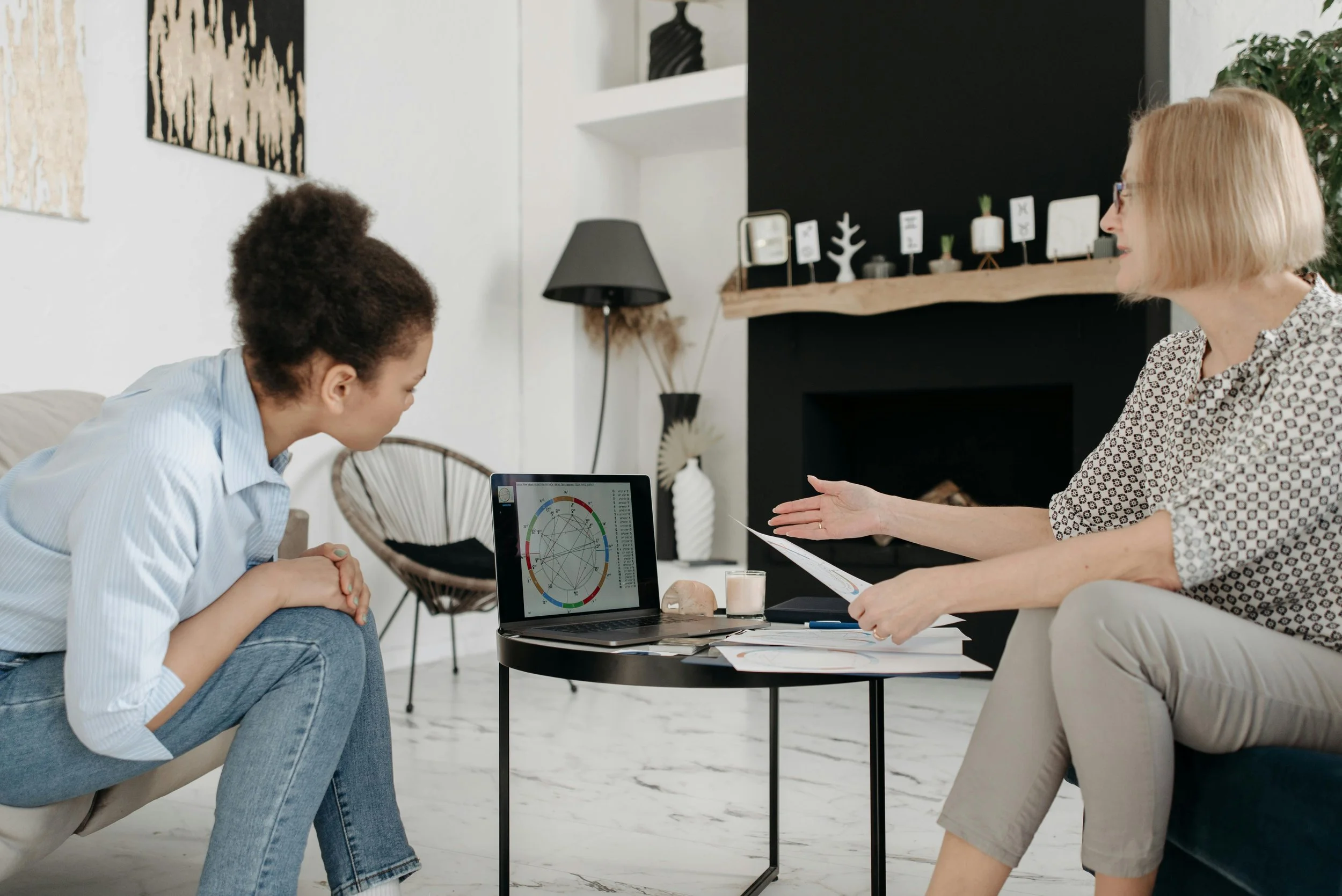 Astrology mentor guiding a student through a live chart reading using a laptop and printed natal charts