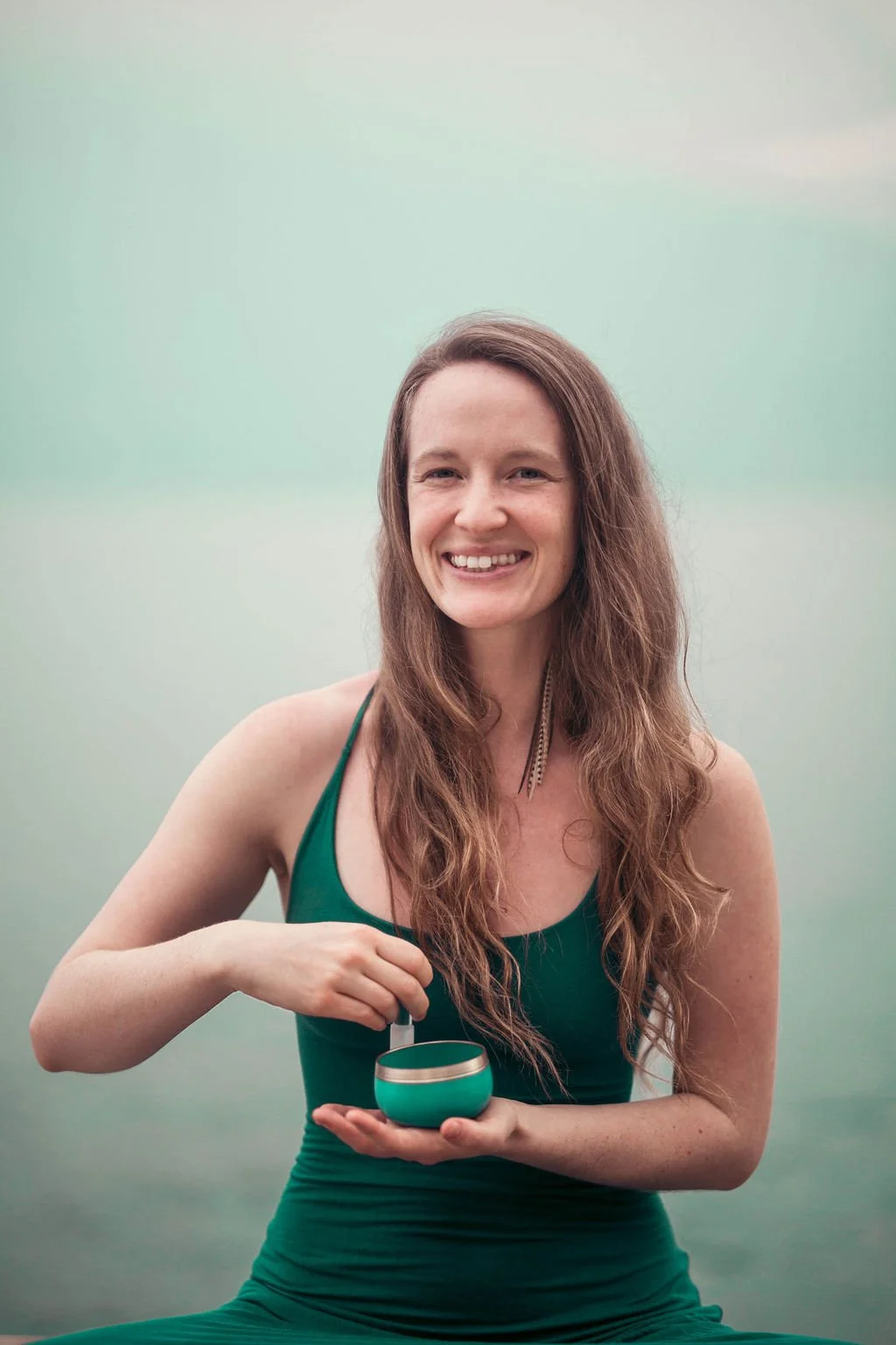 A woman with long wavy brown hair, wearing a green tank top, smiling, holding a singing bowl, sitting outdoors with a misty background guiding others on their path to purpose with Evolutionary Astrology
