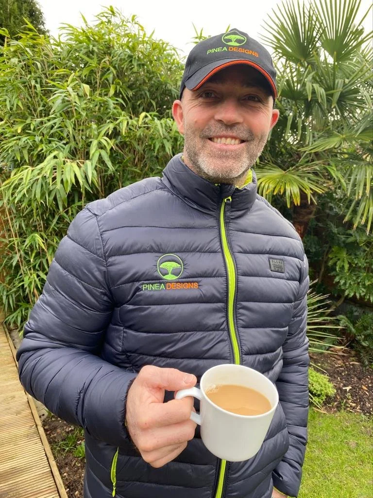Marc Rice holding a mug of coffee outdoors, wearing a black cap and a gray quilted jacket, in front of lush green plants.