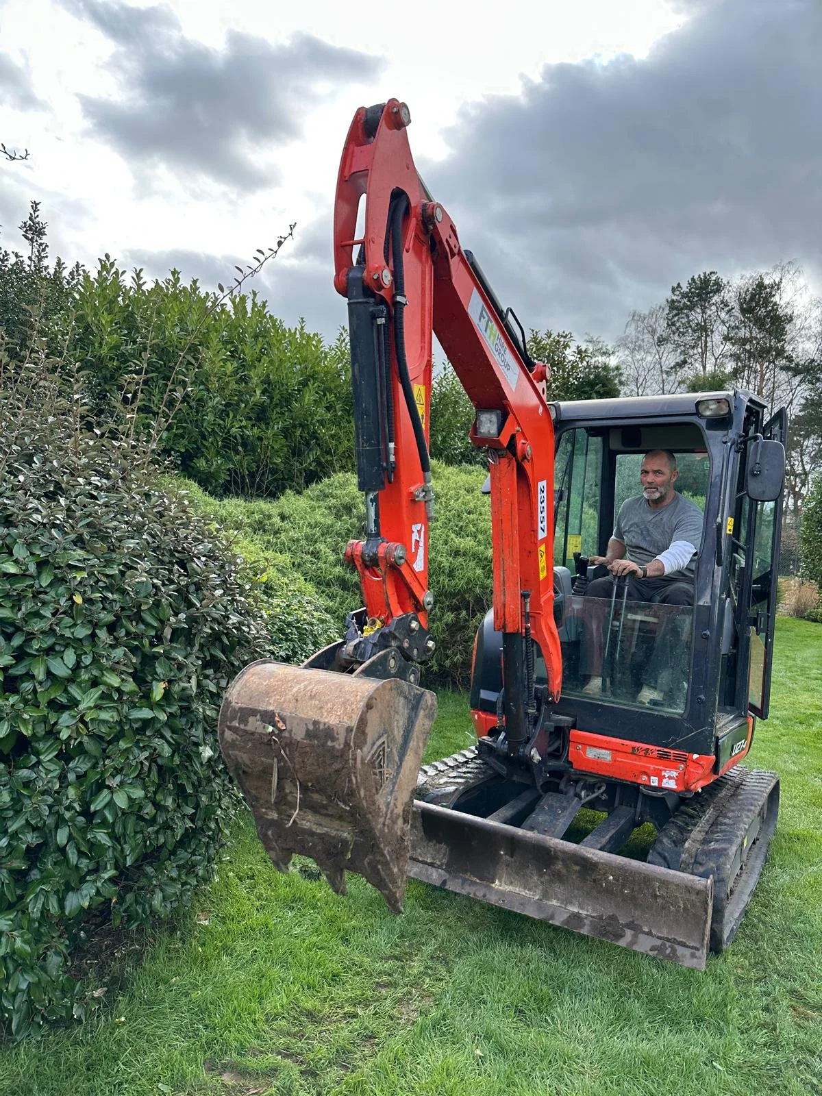 A man (Marc) operating a small red and black excavator, clearing bushes in a grassy area outdoors under cloudy skies.