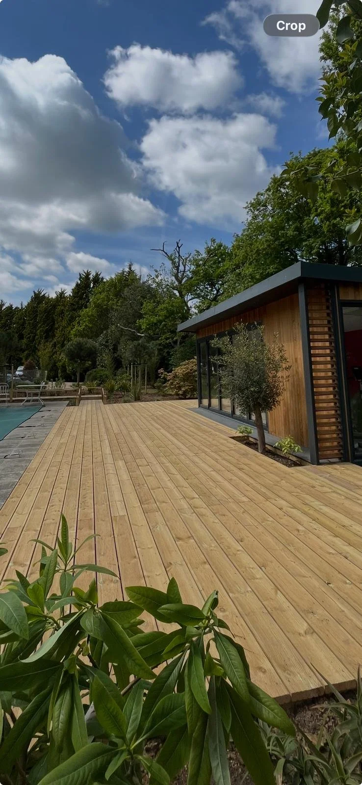 View of a modern wooden house with large glass windows, surrounded by trees, on a wooden deck next to a swimming pool under a partly cloudy sky.