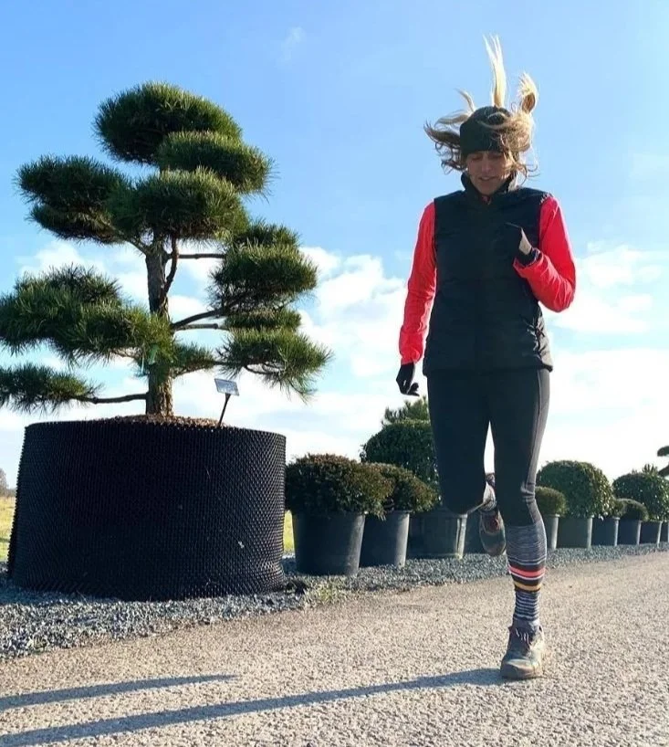 Debbie Boxall jogging outdoors on a sunny day, wearing athletic clothes and striped socks, with potted plants and trees in the background.