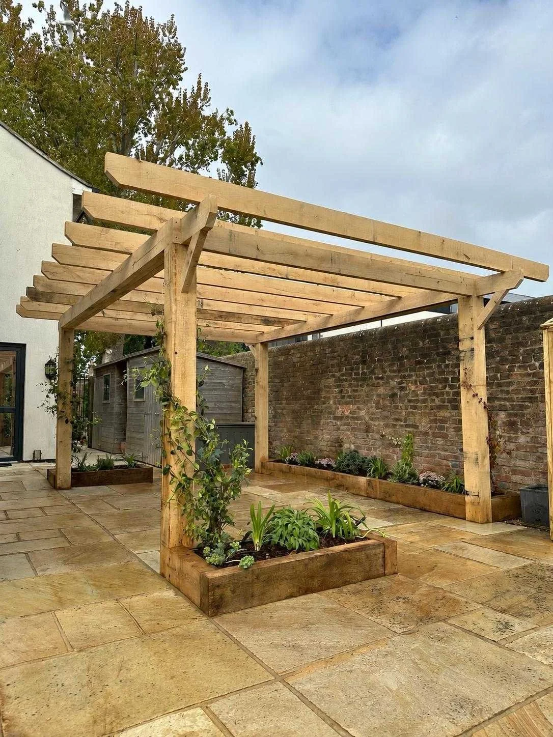 A wooden pergola frame with plant beds on a tiled patio, next to a brick wall and a house.