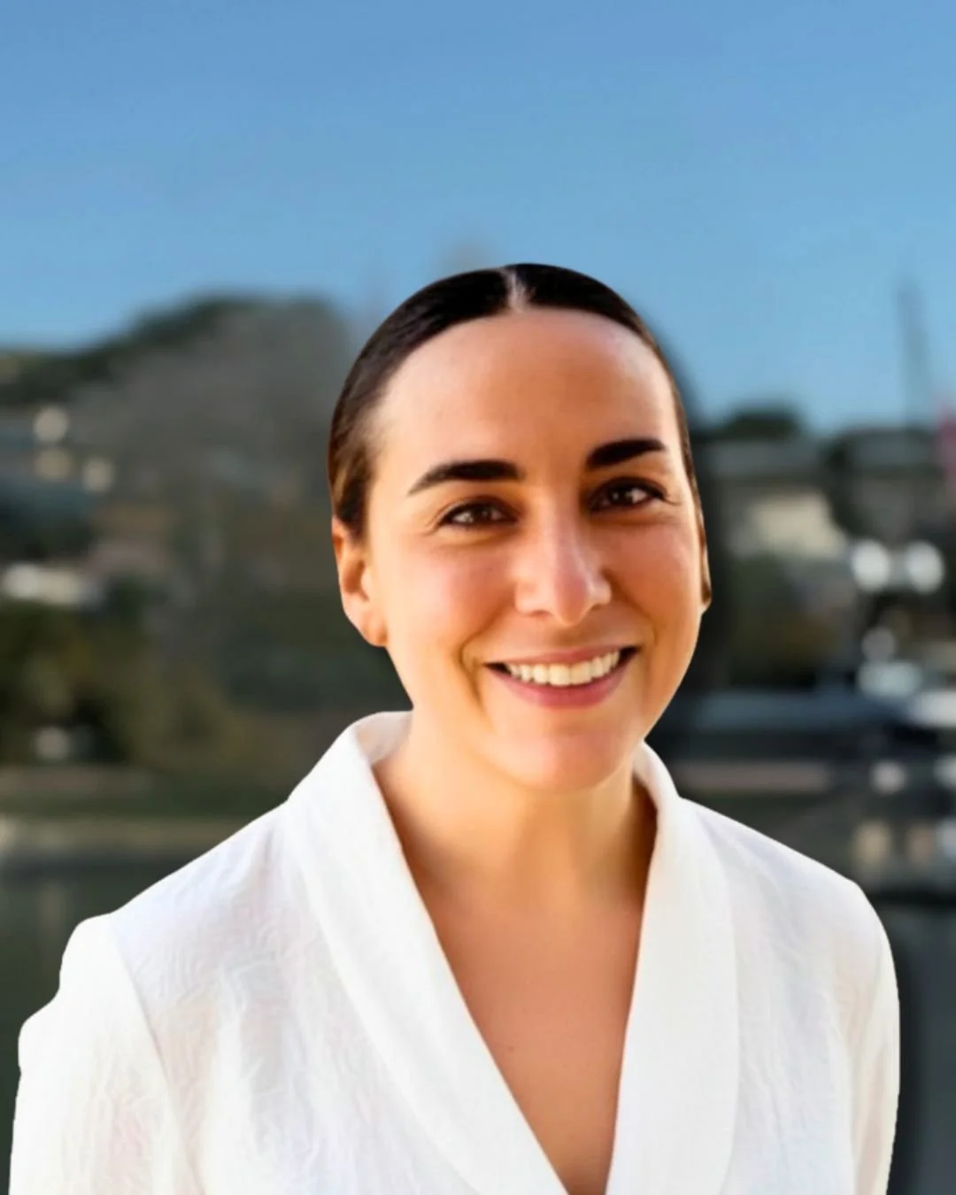 Portrait of a young woman in a white uniform with the logo 'Hatt Mill,' standing outdoors near water with buildings and a hill in the background.