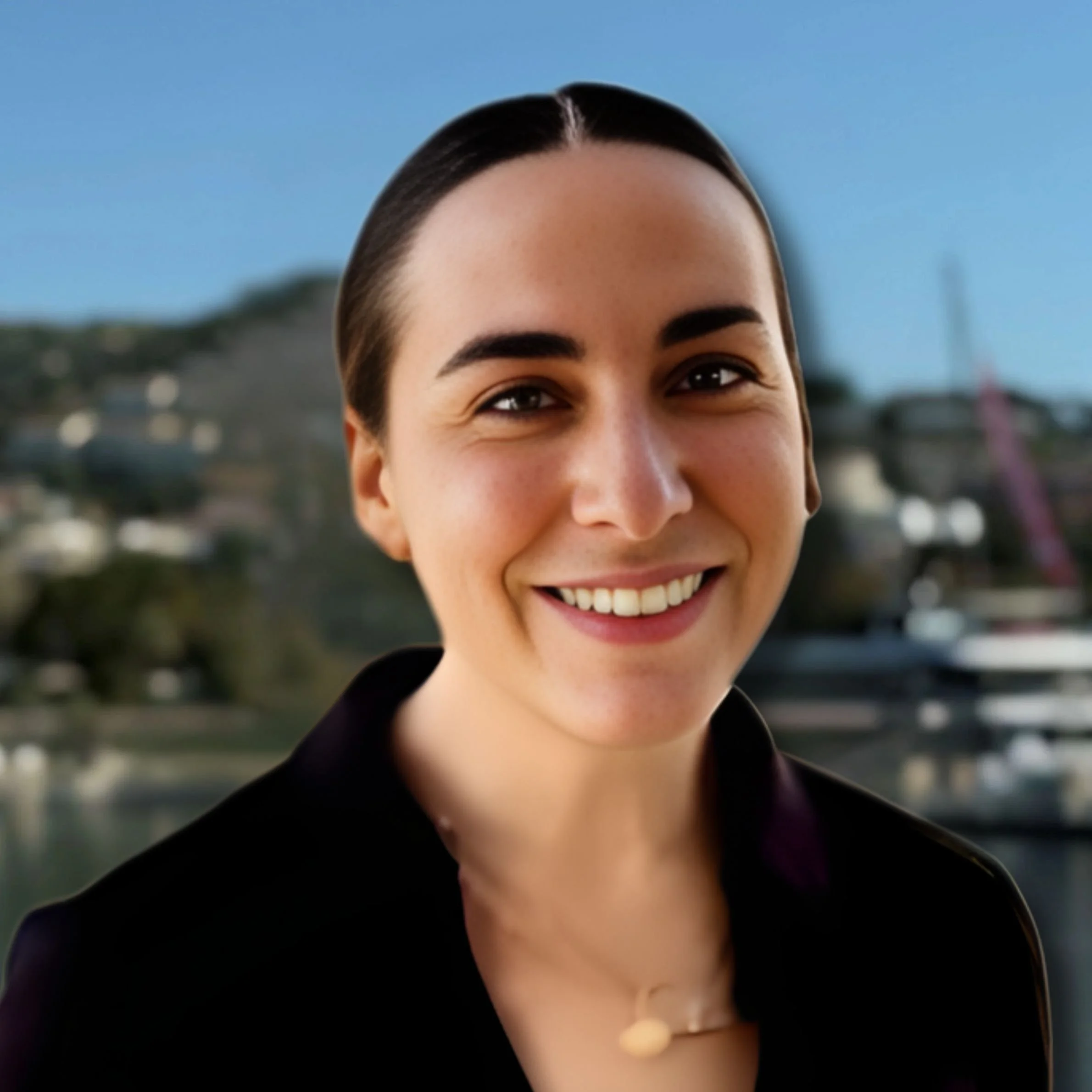 A woman with dark hair smiling at camera with a blurred harbor and buildings in the background.