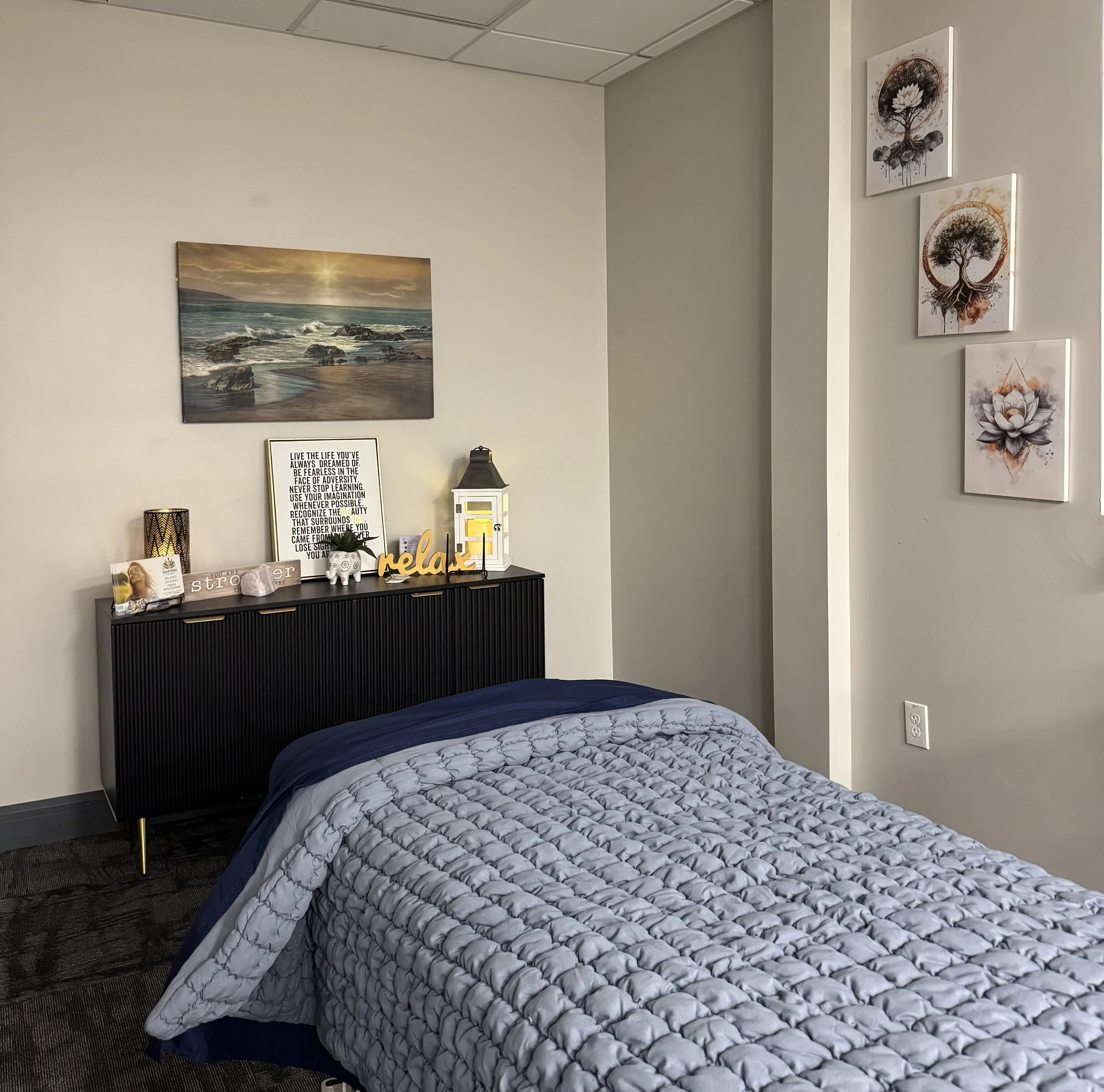 A neatly made bed with a textured gray and blue bedspread in a bedroom with decorative wall art and a black dresser decorated with candles, a picture frame, a sign saying 'relax,' and a lantern. There is a large seascape painting above the dresser and three smaller art pieces featuring trees and a lotus flower on the adjacent wall.