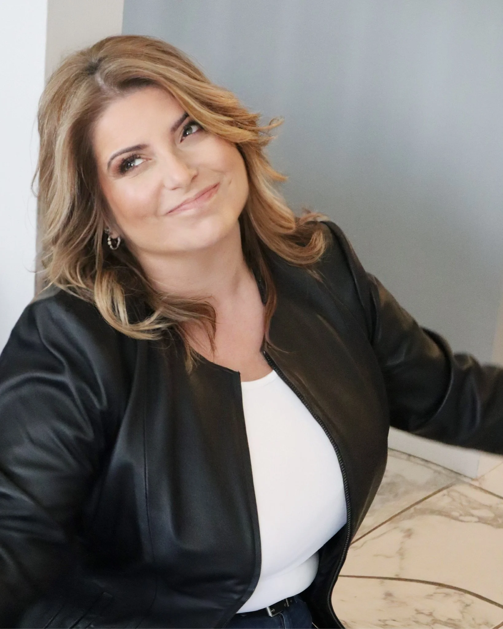 A woman with shoulder-length wavy hair, wearing a black leather jacket and white top, smiling and looking slightly upward.