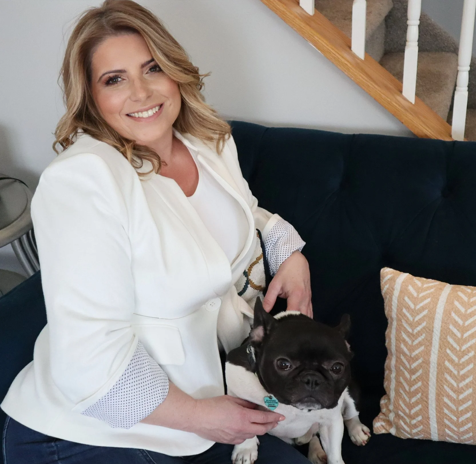 A woman sitting on a dark blue couch, smiling, with a black and white French Bulldog on her lap.