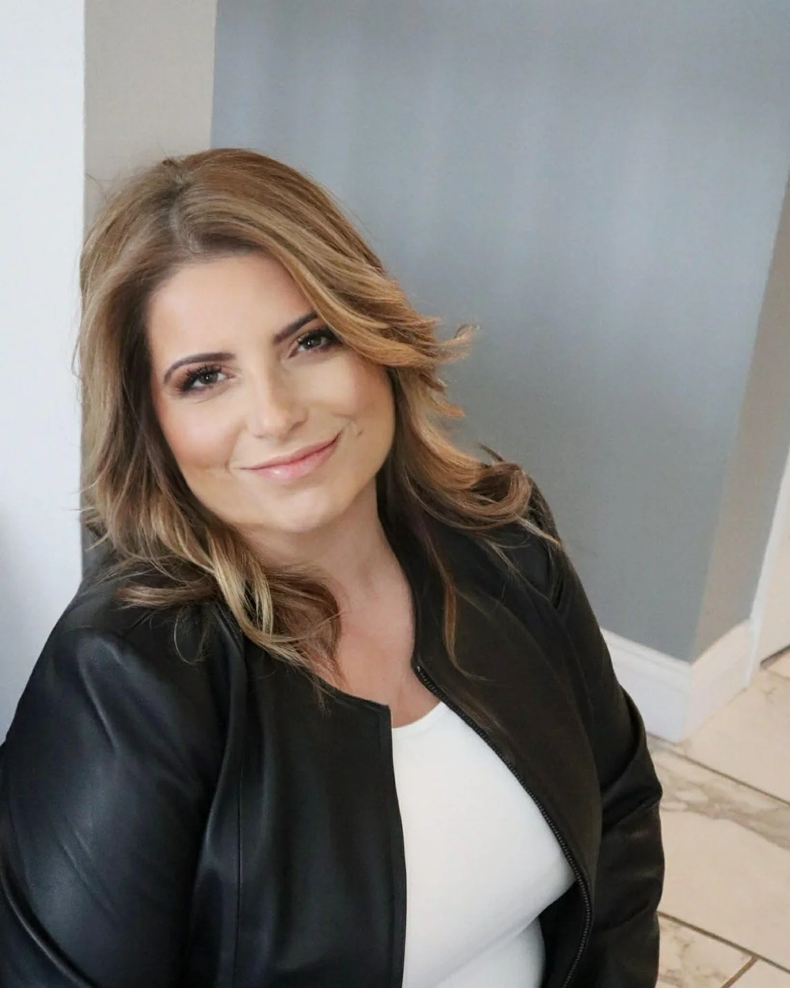 A woman with shoulder-length wavy light brown hair smiling, wearing a black leather jacket over a white top, and posing indoors against a light-colored wall.