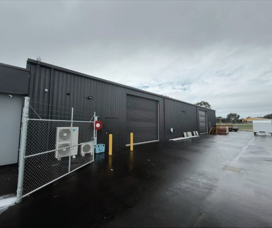 Industrial building with black metal exterior, chain-link fence with air conditioning units, yellow safety posts, and equipment containers on wet asphalt ground under a cloudy sky.