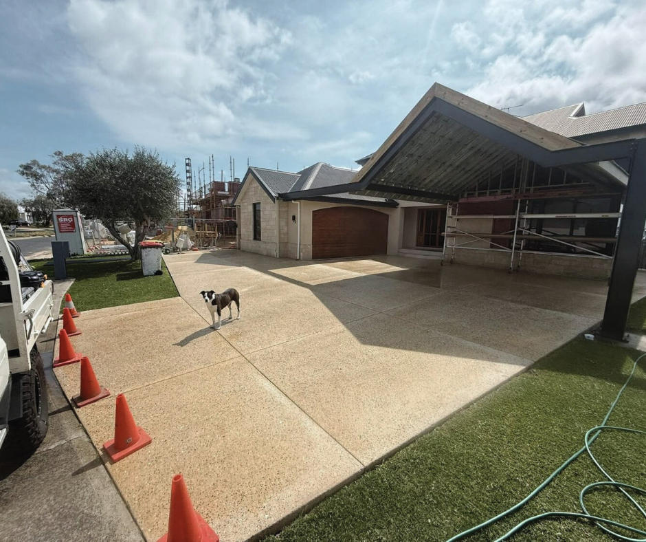 Newly constructed house with a driveway, a black and white dog standing on it, construction scaffolding and equipment visible, with a tree, grass, and cloudy sky in the background.