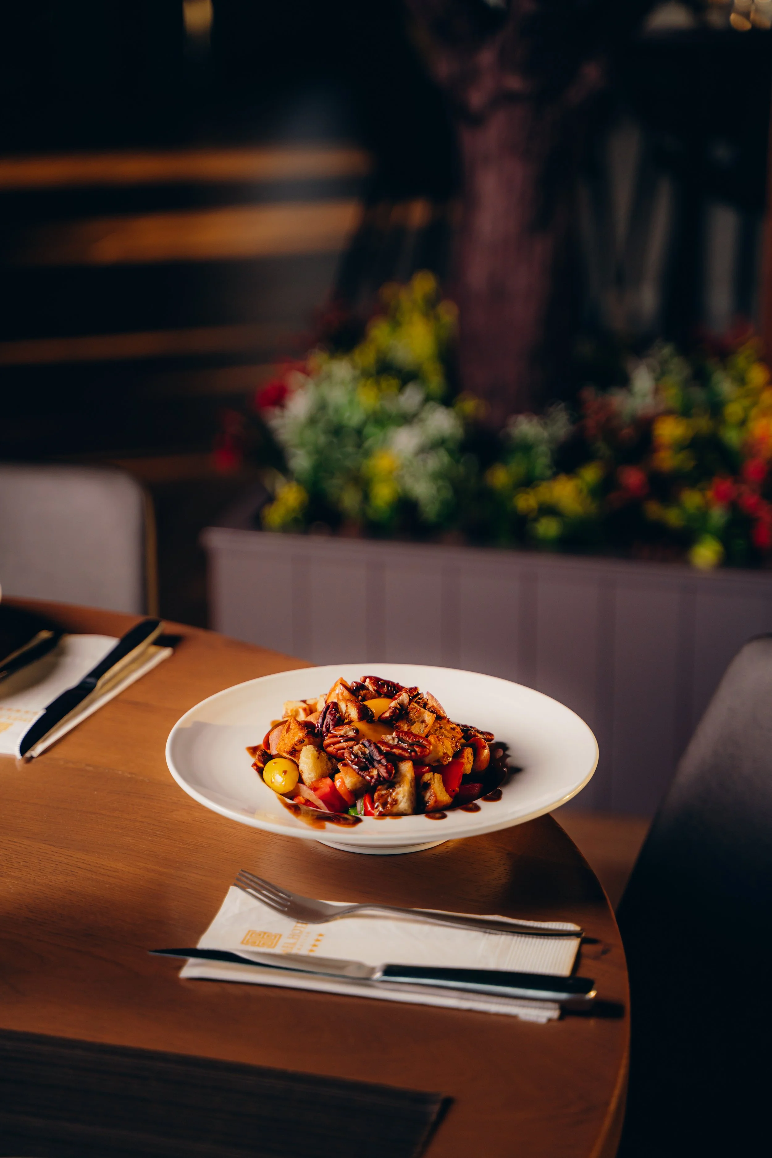 A plate of food with mixed ingredients including vegetables, nuts, and sauce, on a wooden table, with utensils and a napkin nearby, in a dimly lit restaurant setting with decorated walls.