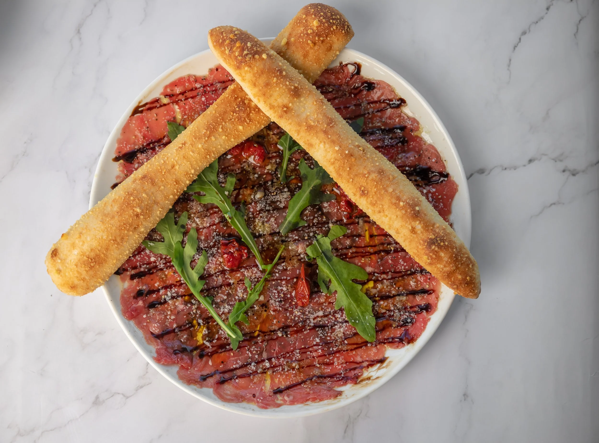 A white bowl filled with carpaccio topped with arugula, balsamic glaze, and grated cheese, garnished with three breaded cheese sticks.