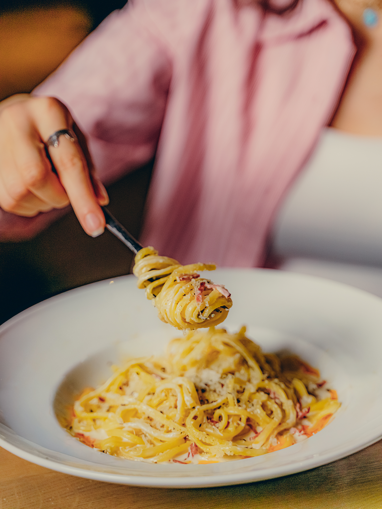 Close-up of a bowl of spaghetti pasta with bacon and cheese, with a person in a pink sweater using a fork to pick up a strand of pasta.