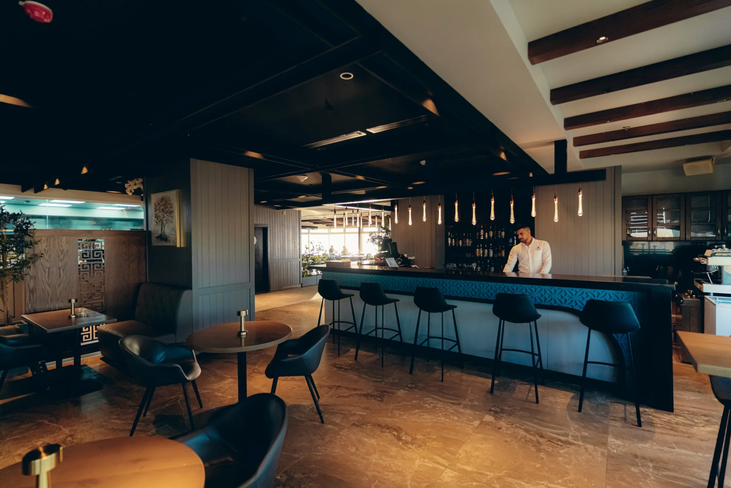 Interior of a modern restaurant with a bar counter, black chairs, and a bartender preparing drinks. The ceiling has wooden beams and hanging decorative lights. There are large windows in the background letting in natural light.