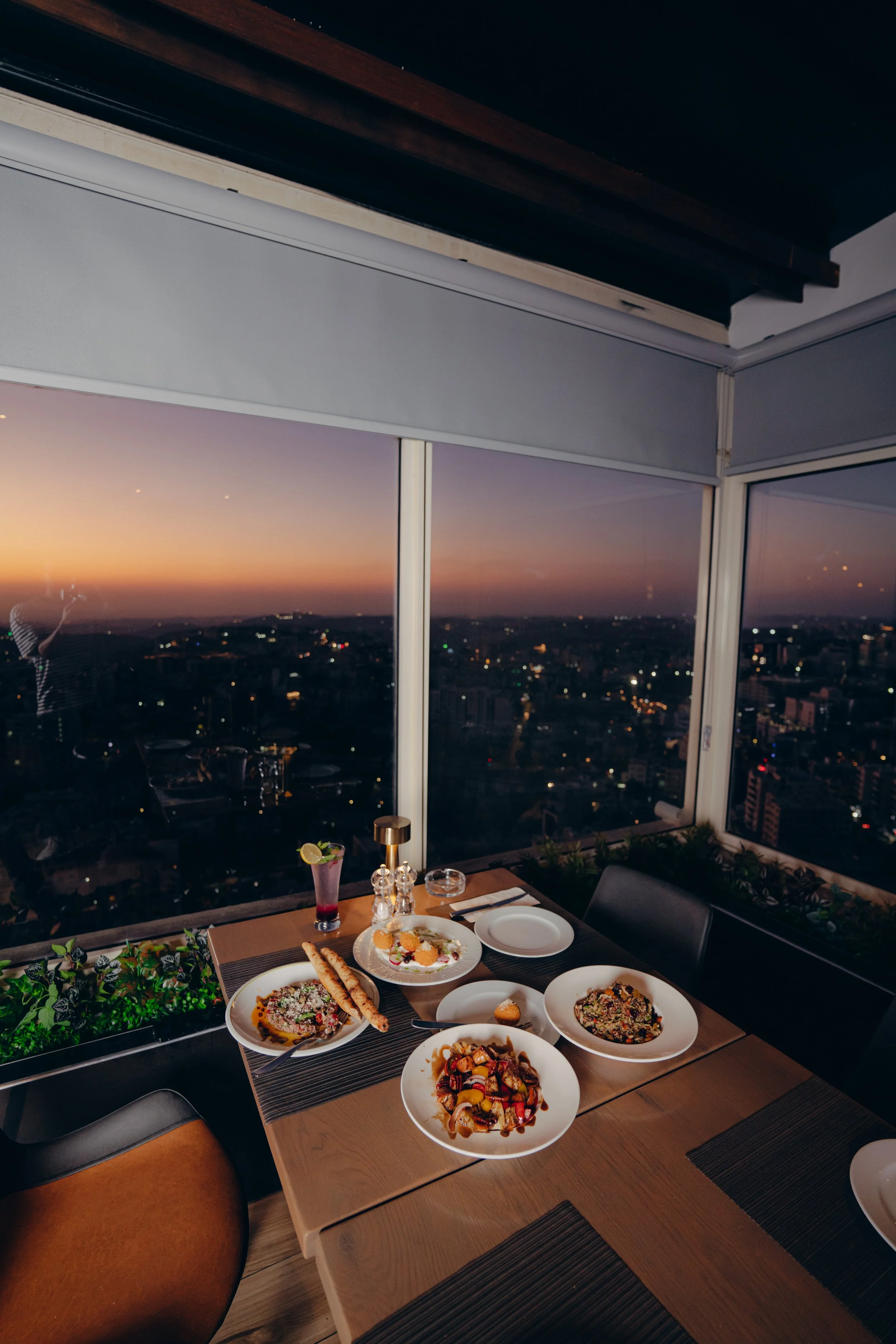 A dining table set with various plates of food, drinks, and utensils in a modern restaurant with a city view at sunset.