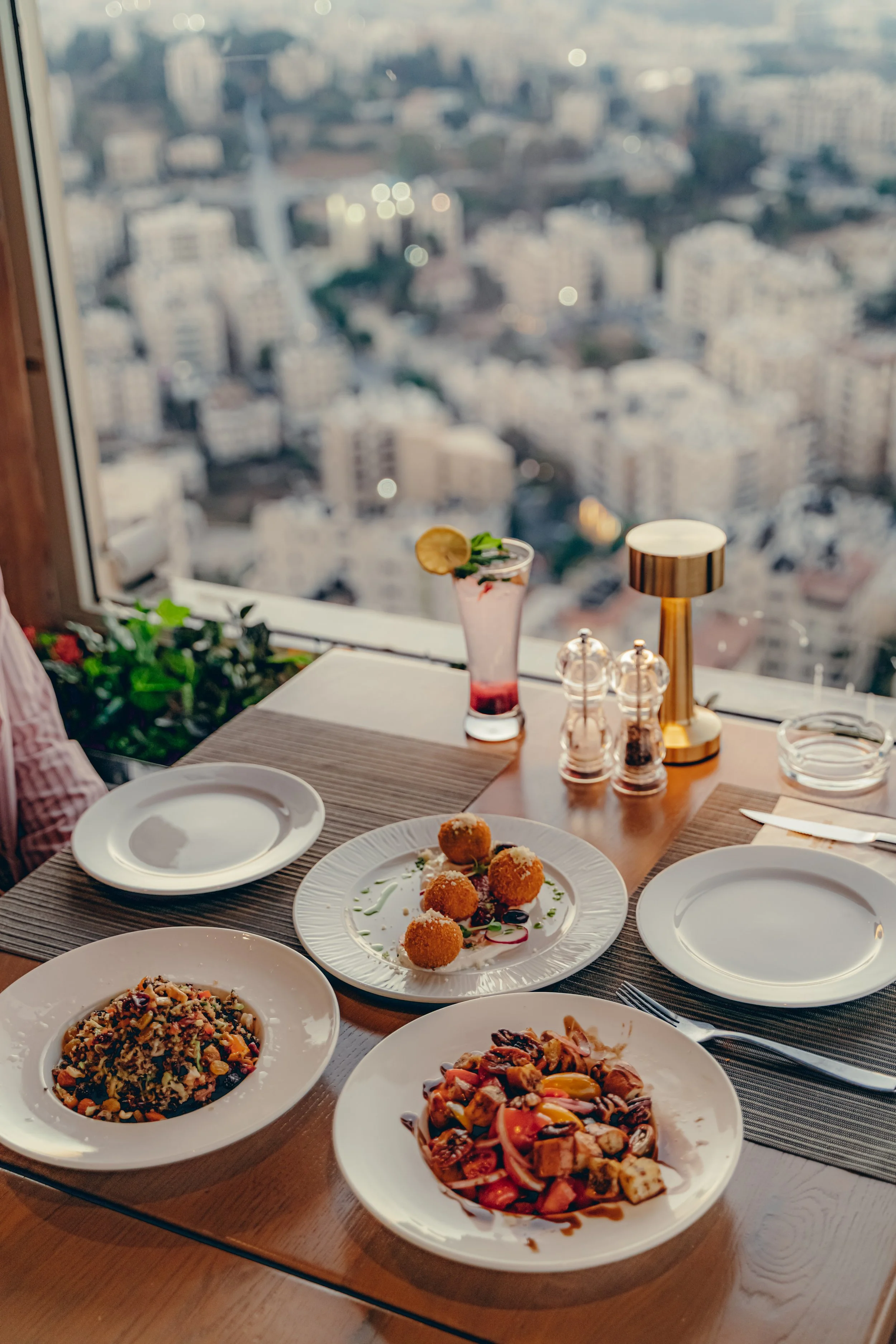 A dining table with various dishes, including fried appetizers, pasta, and salad, set in front of a large window overlooking a cityscape with tall buildings.