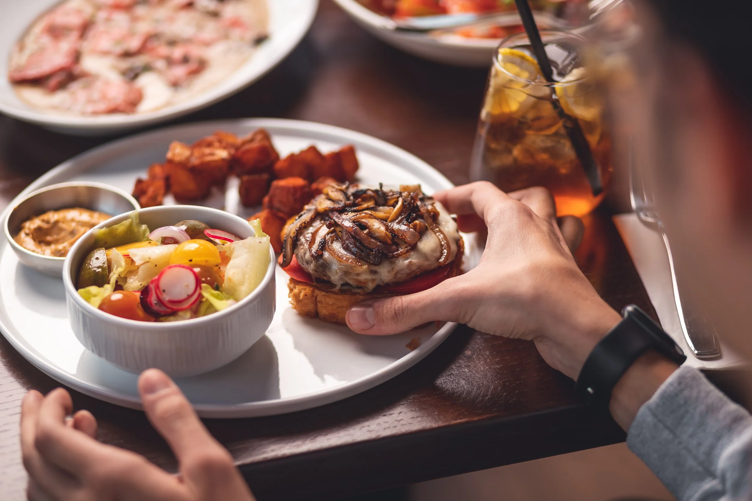 A person holding a tray with a burger topped with mushrooms and onions, a bowl of salad with radishes, cherry tomatoes, and pickles, a small dish of sauce, and a plate of fried chicken pieces, with other dishes and a glass of iced tea in the background.