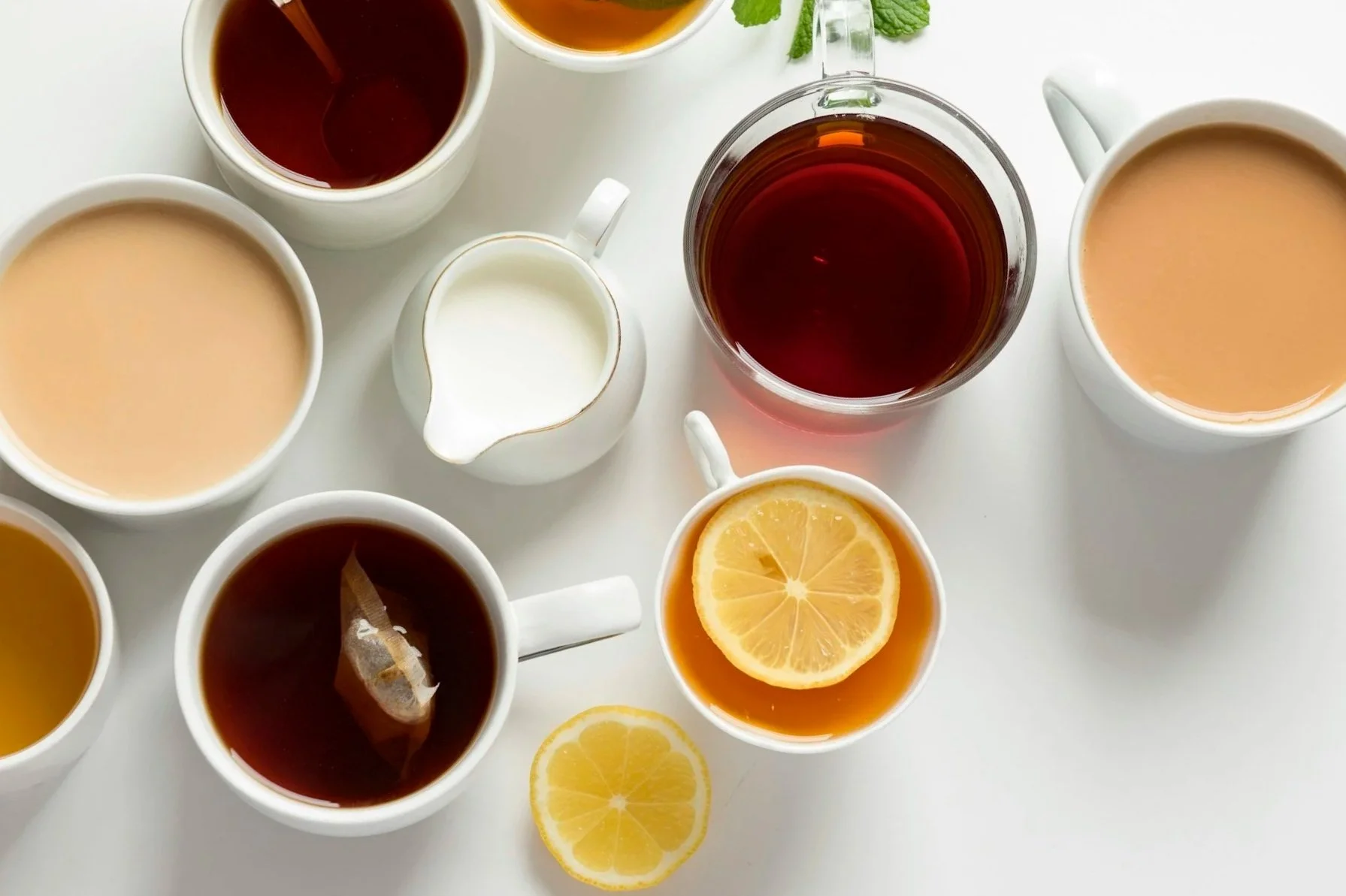 Multiple cups of tea, a glass teapot, a small pitcher of cream, and lemon slices are arranged on a white surface. Some tea is dark, and some is light.