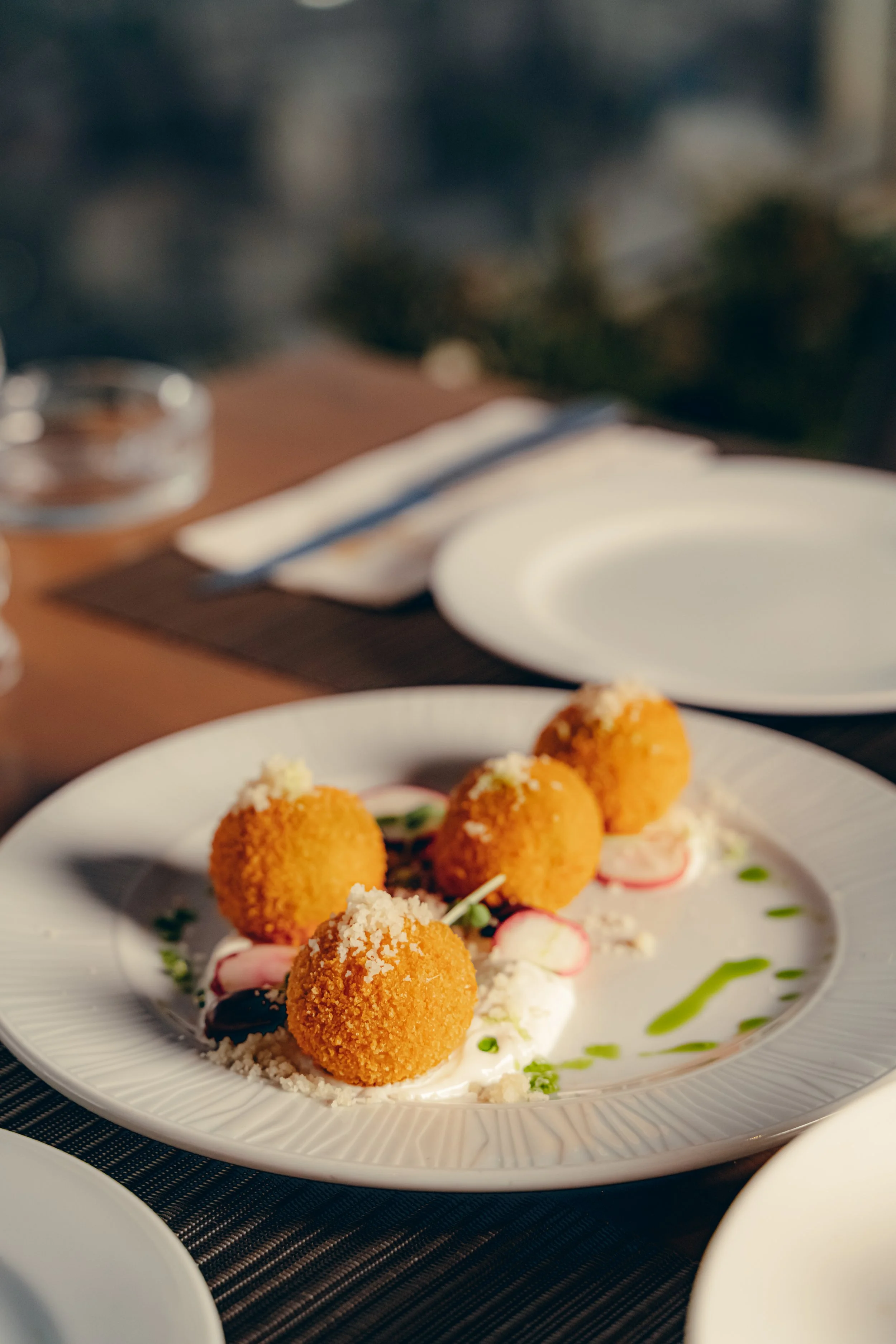 Close-up of a white plate with four golden, crispy fried balls garnished with grated cheese, microgreens, and thin slices of radish, drizzled with green sauce, on a dark table. In the blurry background, there is a clear glass of water, a fork wrapped in a napkin, and an empty white plate.