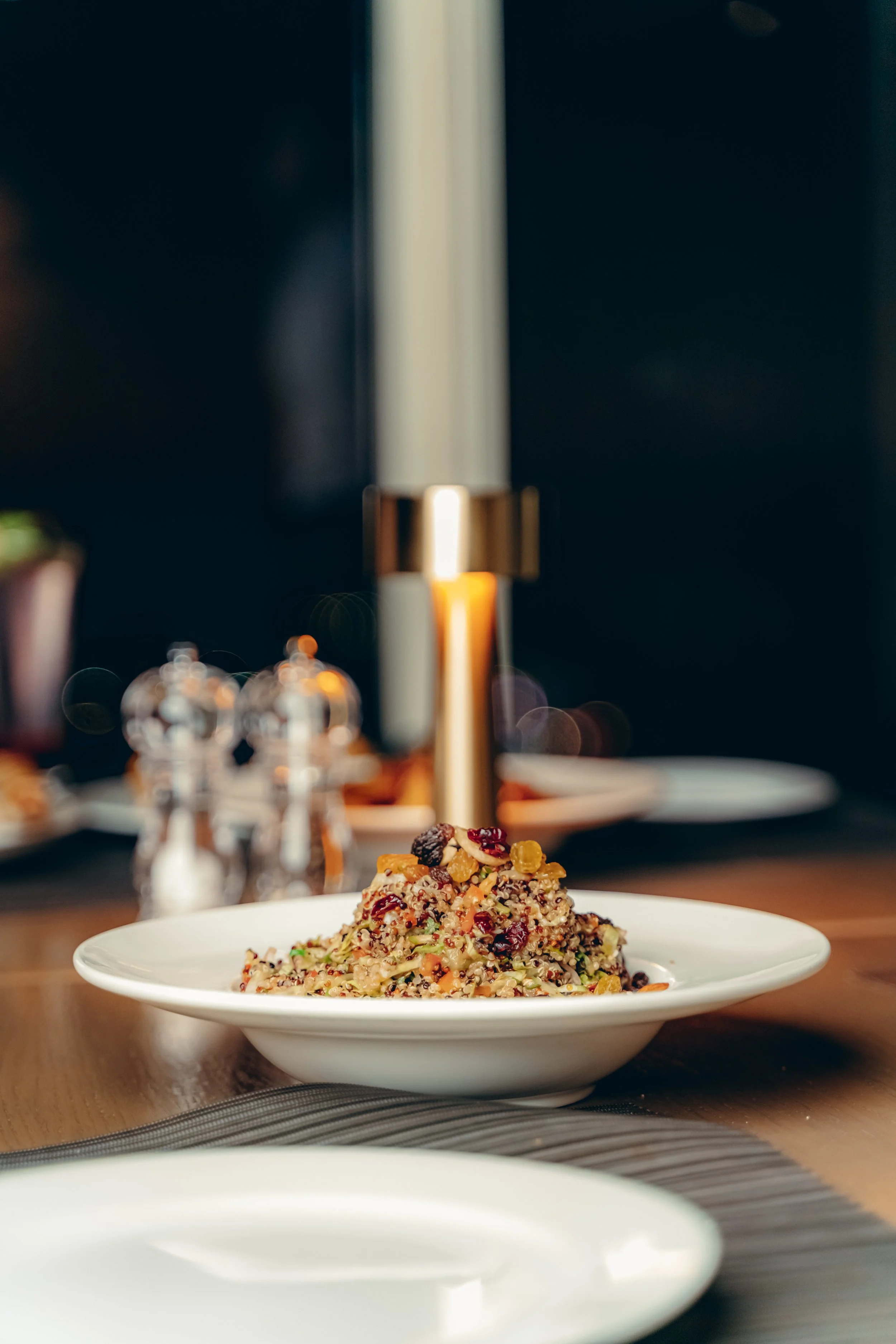 A plate of quinoa salad with vegetables and dried fruit on a wooden table, with a blurred background of a modern kitchen or restaurant setting.