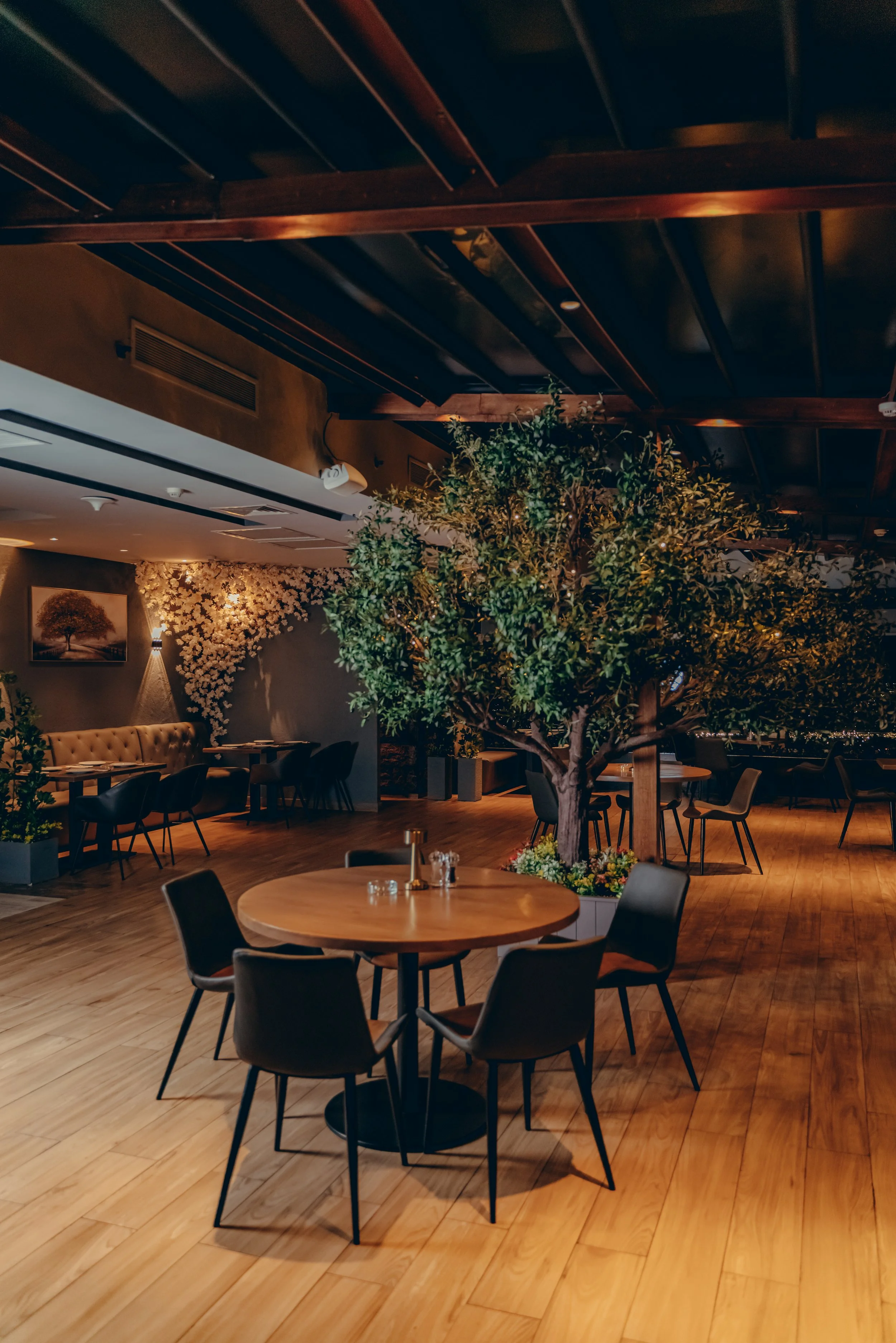 Interior view of a restaurant with wooden flooring, round tables with black chairs, a large potted tree, and decorative floral wall art.