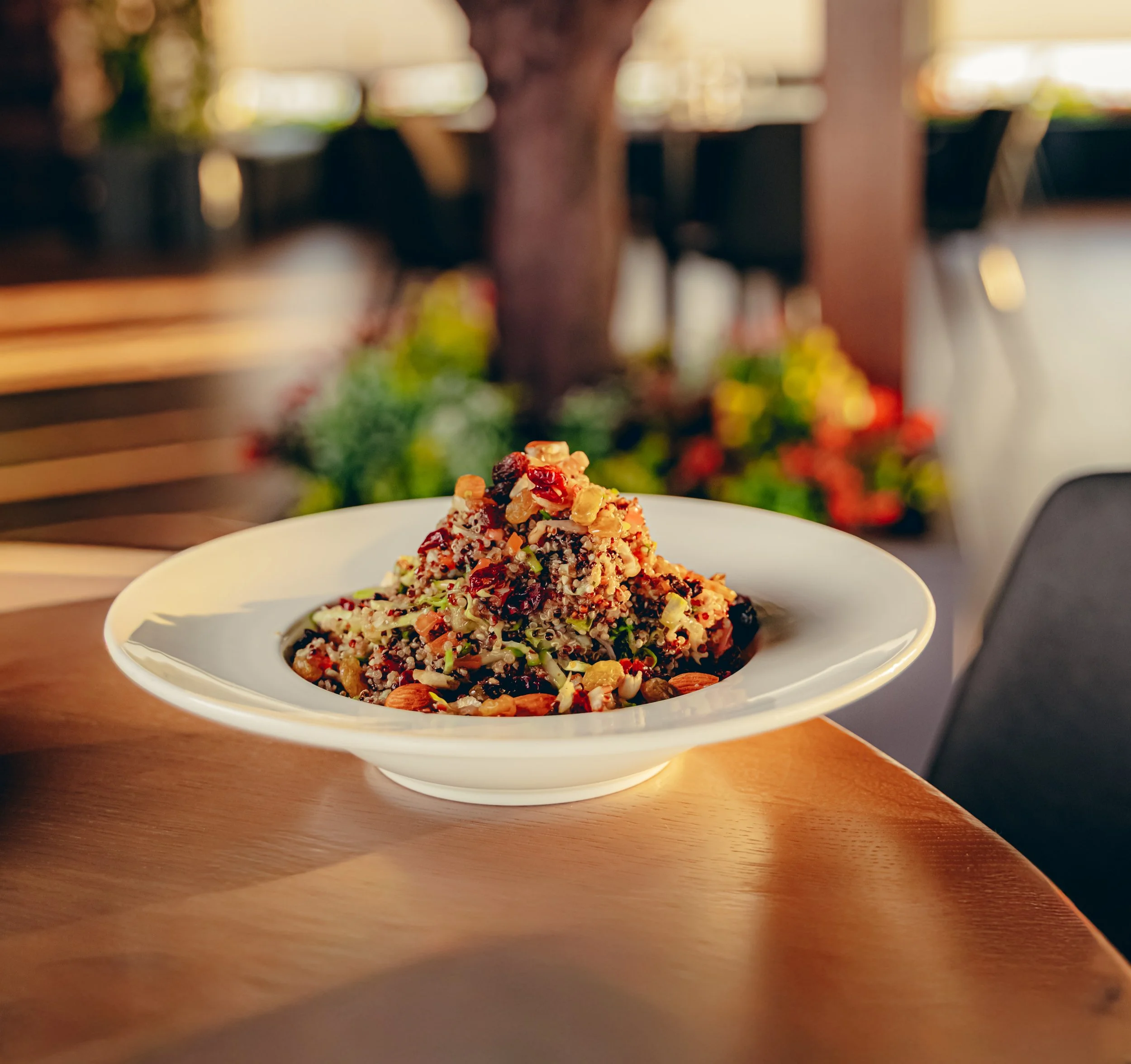 A close-up view of a plated dish with a small serving of food, which appears to be a mixture of grains, nuts, and dried berries, placed on a light-colored plate on a wooden table. The background is blurred with warm lighting and some greenery and colorful objects.