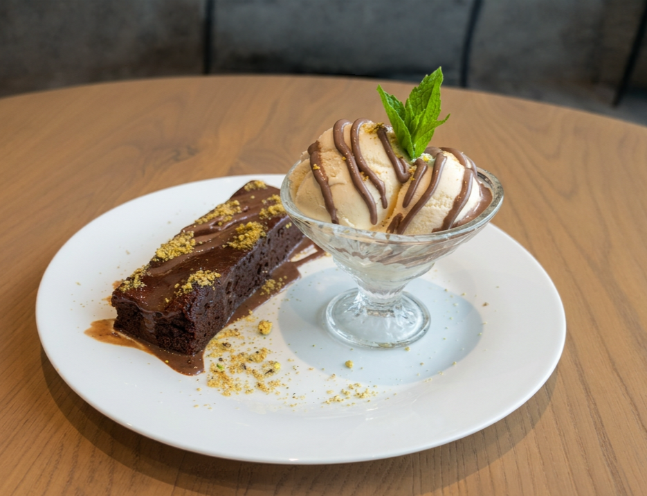 Chocolate brownie and vanilla ice cream topped with chocolate syrup and a mint leaf on a white plate.