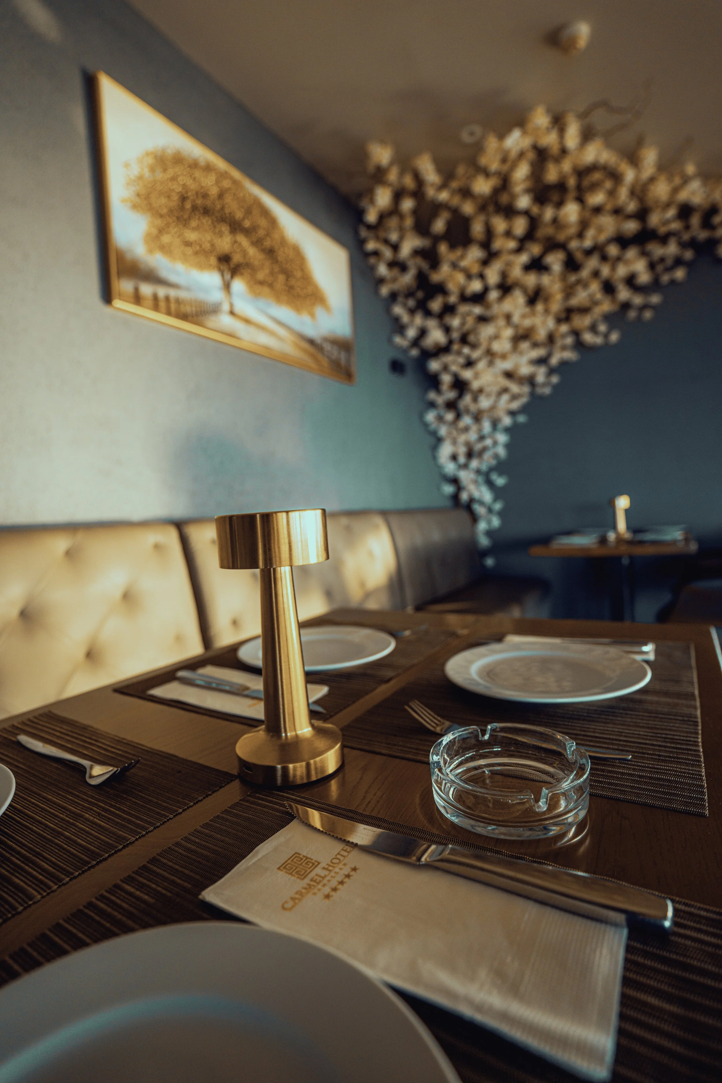 Elegant restaurant table setting with plates, cutlery, a glass ashtray, a gold table lamp, and a menu with a hotel logo in a cozy dining room decorated with framed artwork and a floral installation on the wall.