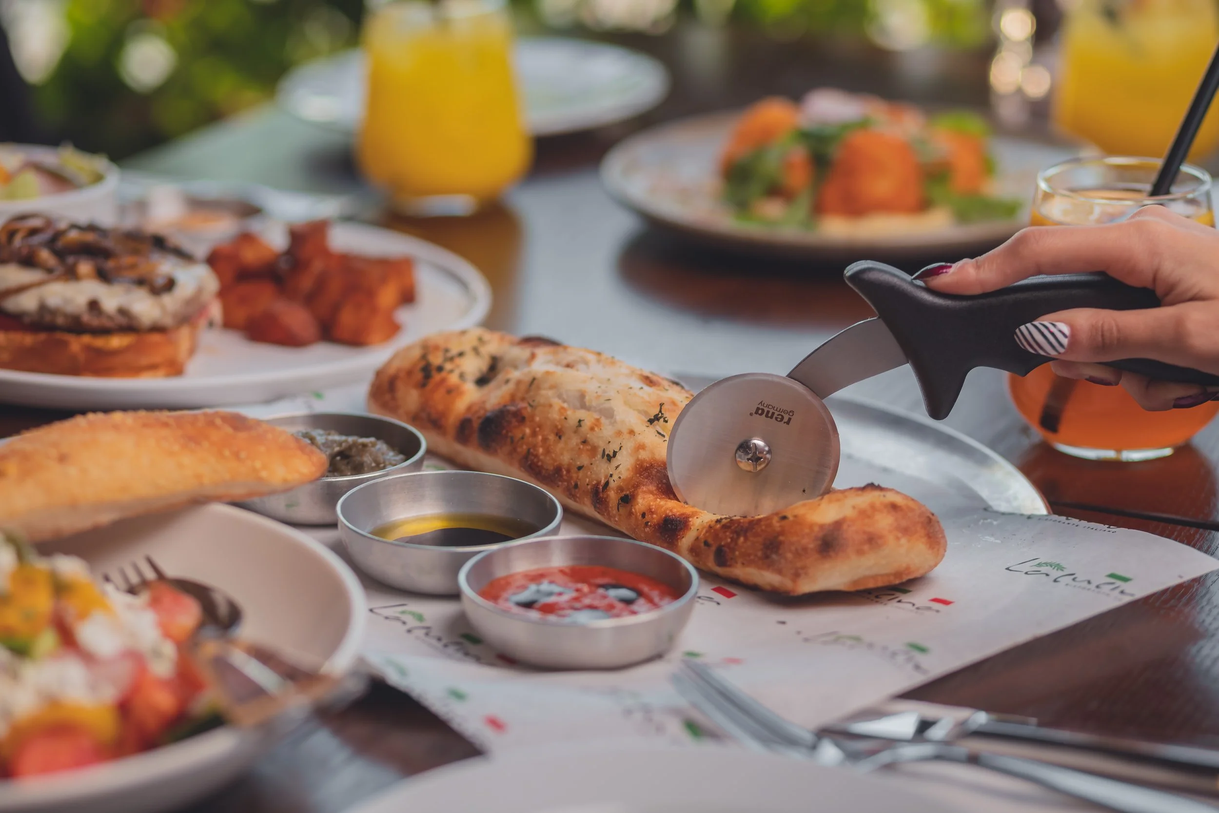A table with various plates of food, including a pizza, chicken wings, and breadsticks, along with small bowls of sauces and drinks, with a person's hand using a slicer to cut a pizza.