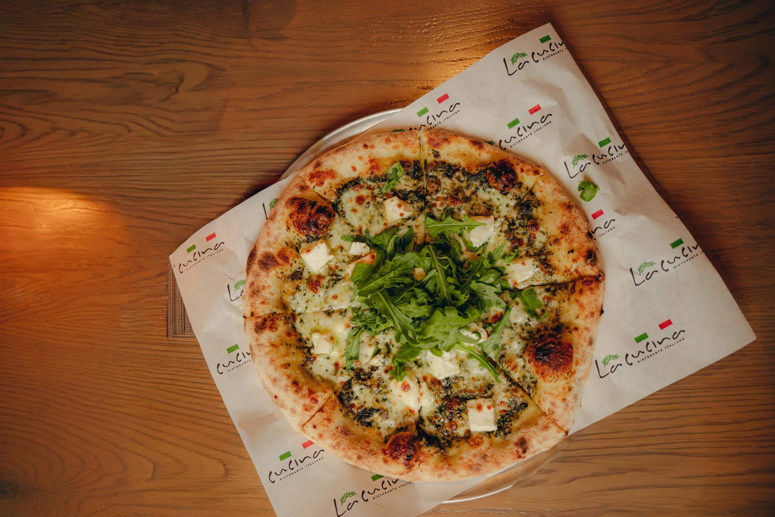 A pizza with cheese, greens, and herbs on a wooden table, partly wrapped in branded paper from La Cucina restaurant.