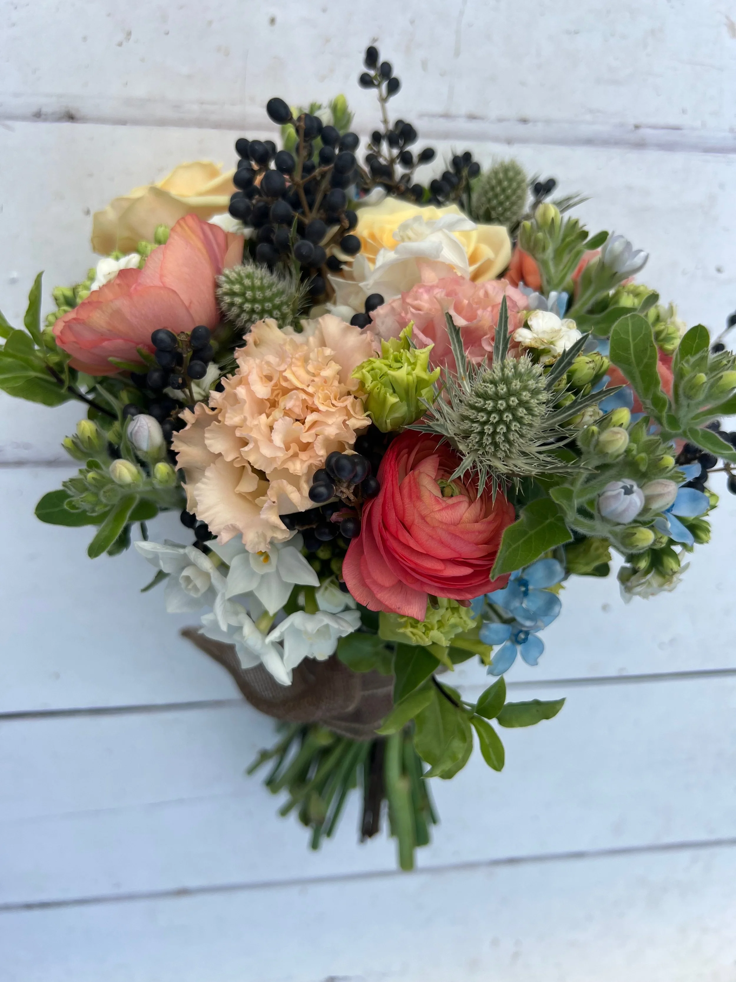 Colorful bouquet of flowers including peach, pink, white, and blue flowers, with green leaves and black berries, arranged in a rustic setting.