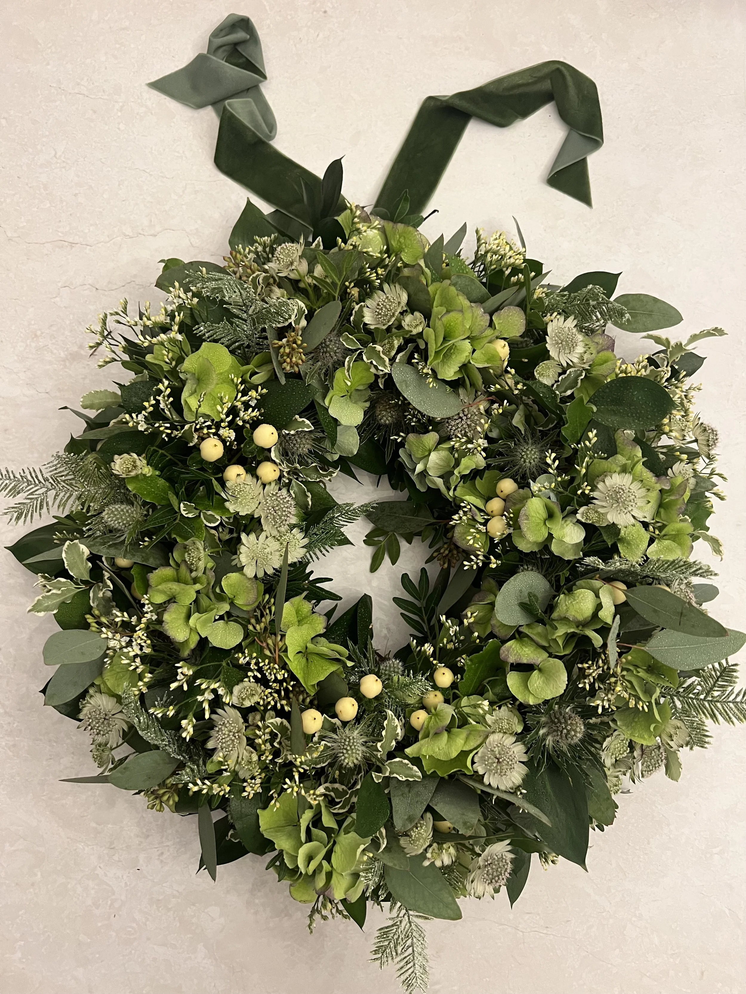 A decorative green wreath made of various green leaves and white flowers, hanging on a light-colored wall, with a green ribbon bow at the top.