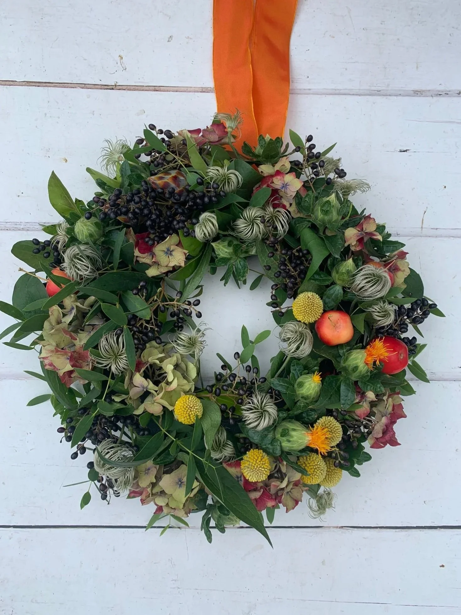 A decorative autumn wreath with green leaves, yellow and red flowers, small apples, black berries, and an orange ribbon, hanging on a white wooden background.