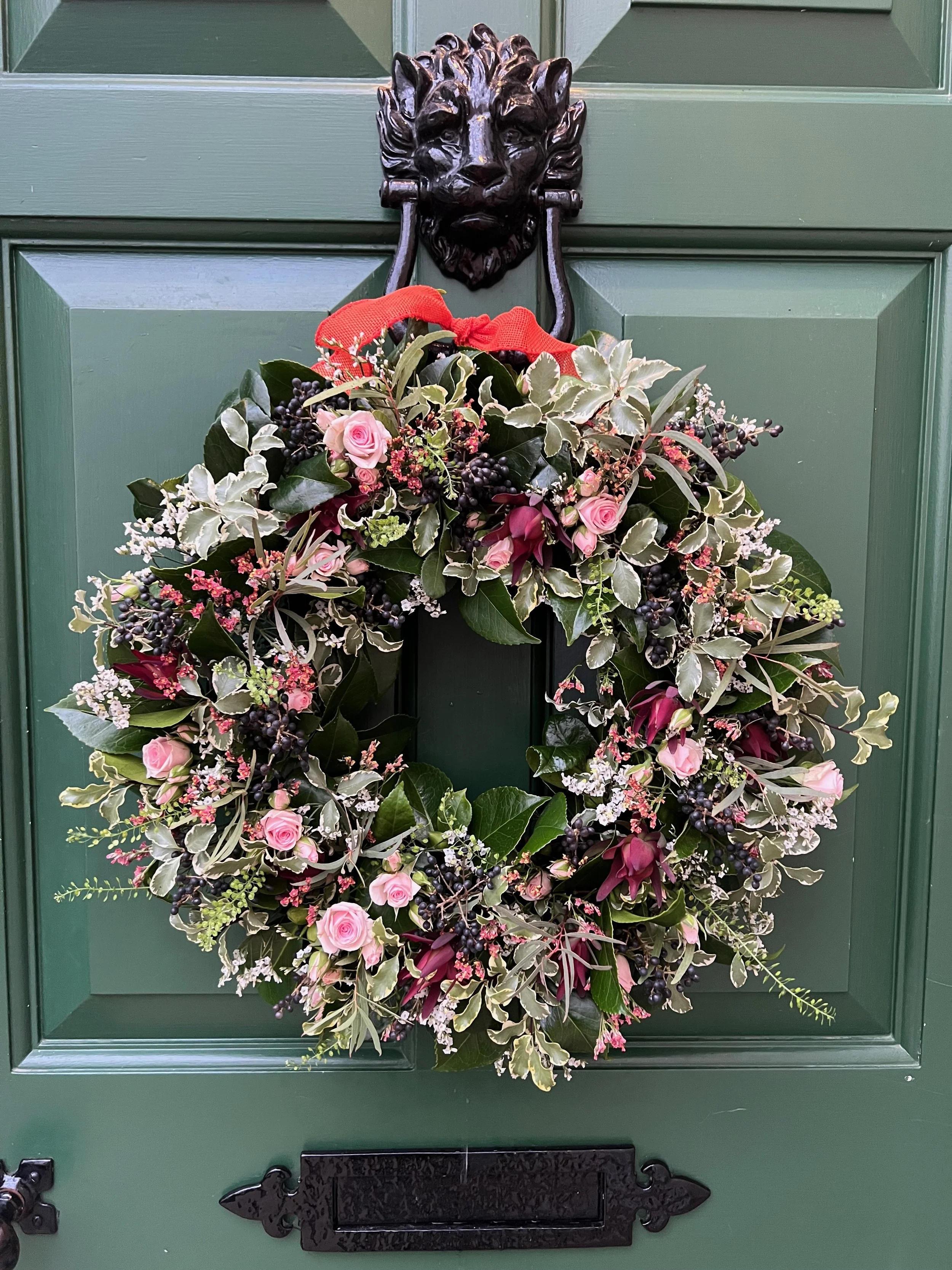 A festive wreath with pink roses, dark berries, and greenery hanging on a green door, decorated with a red bow.
