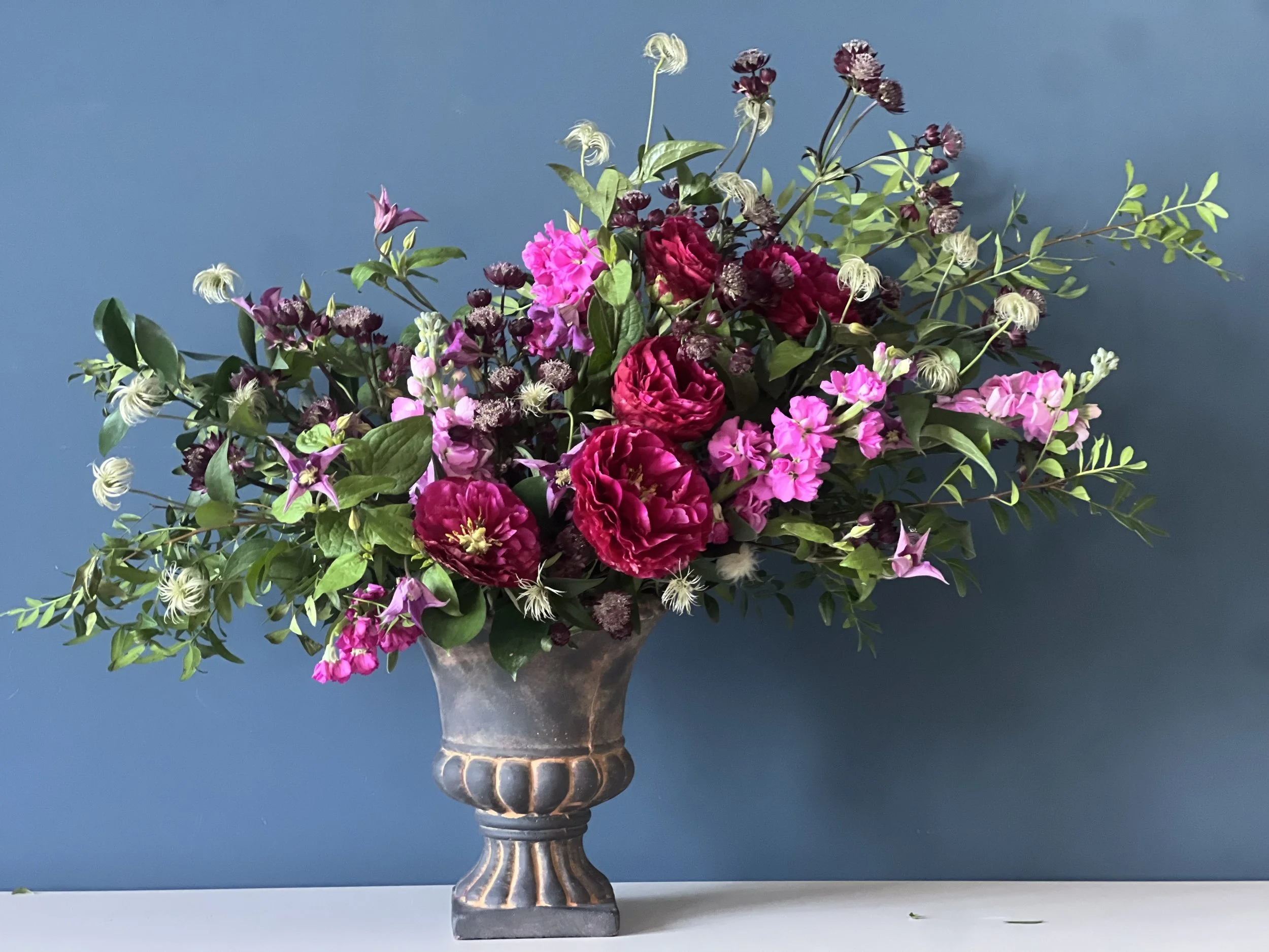 A colorful bouquet of pink, purple, and dark red flowers with green foliage in an ornate gray vase set against a plain blue background.