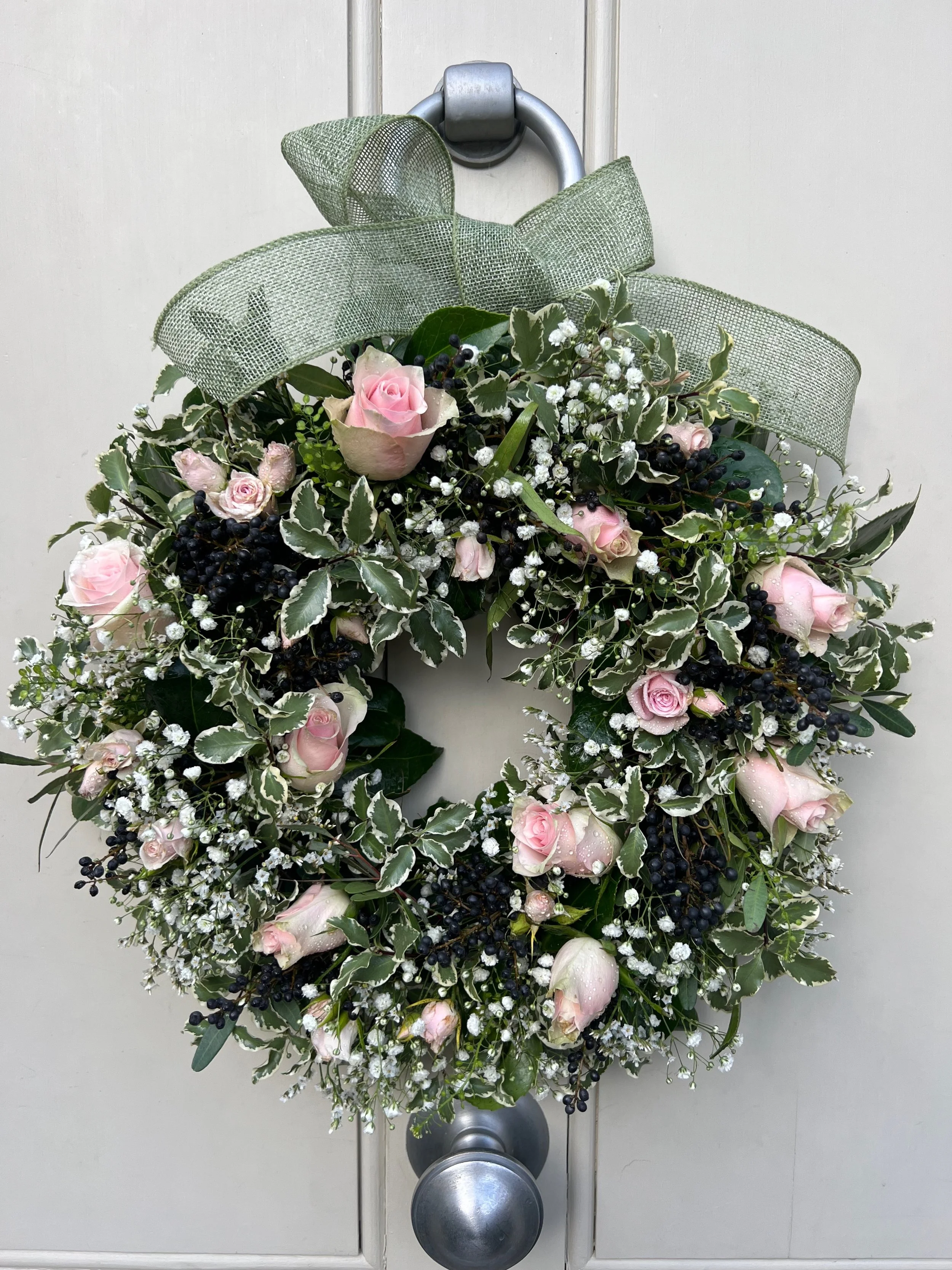 A decorative wreath with pink roses, white baby's breath, and variegated green leaves, adorned with a green mesh bow, hanging on a door knob.