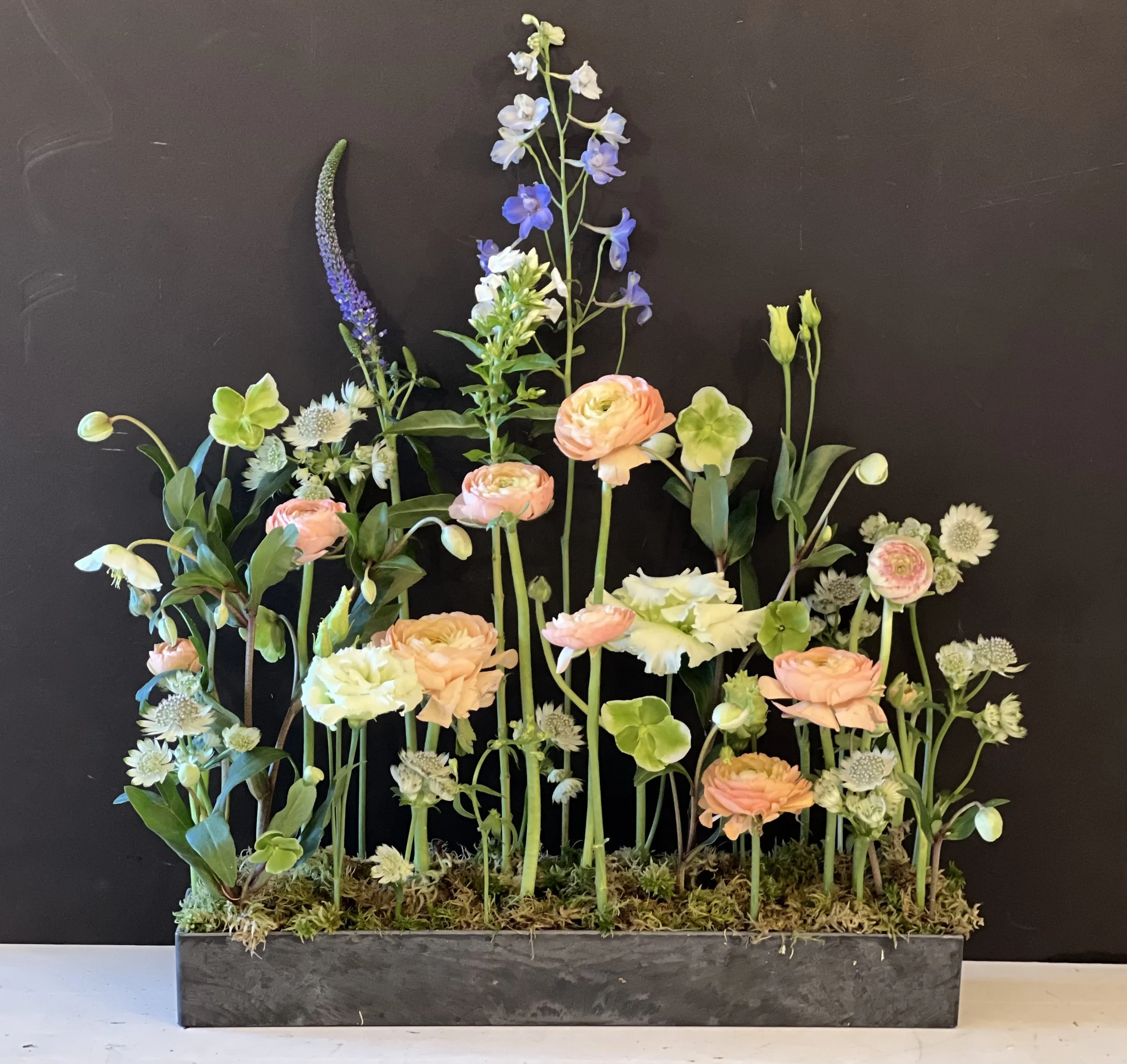 Arrangement of various colorful flowers including ranunculus, delphinium, and others in a rectangular metal container with moss on top, set against a black background.