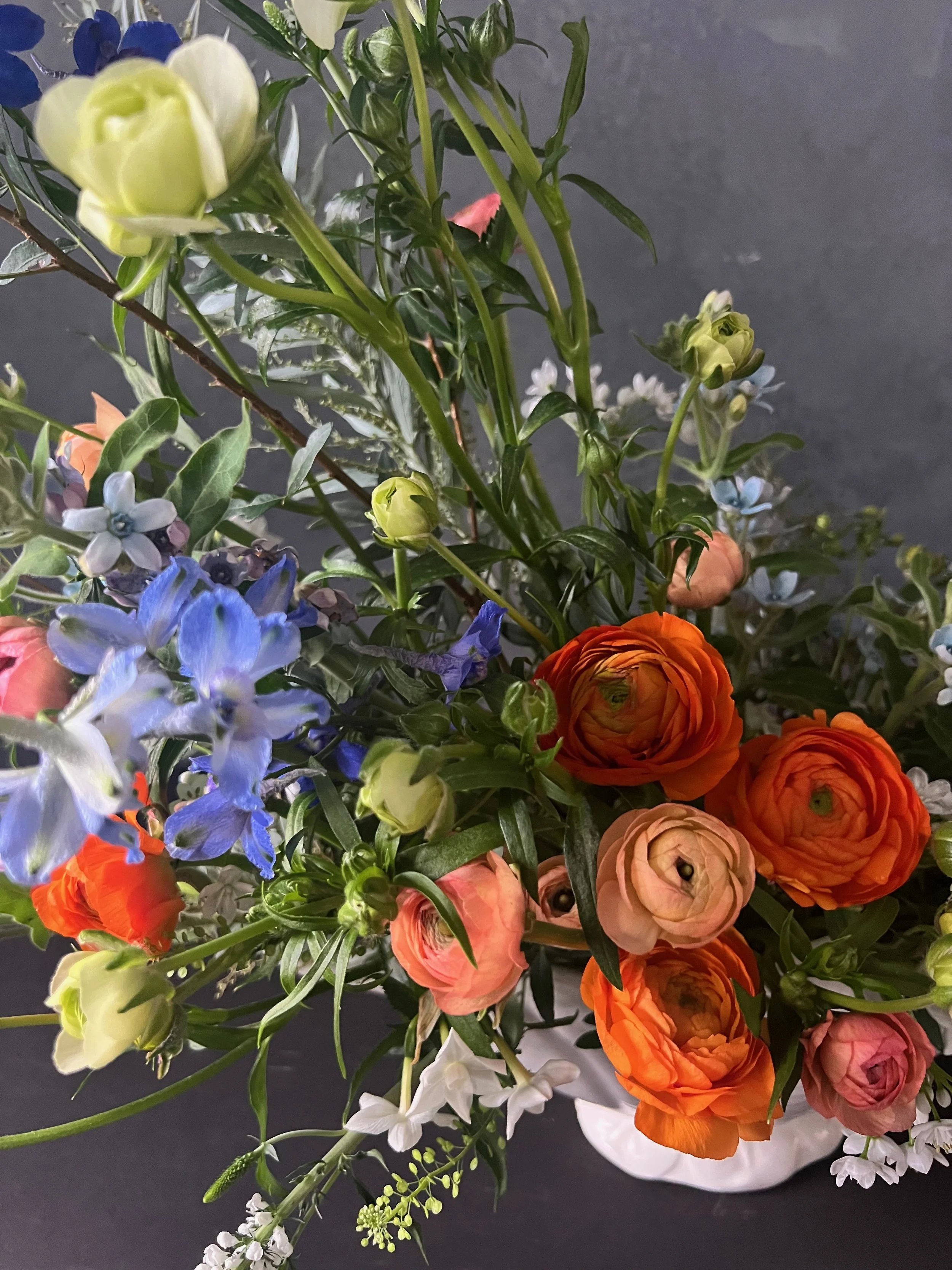 A vibrant flower arrangement in a white vase featuring orange, pink, blue, white, and green flowers and foliage on a dark background.