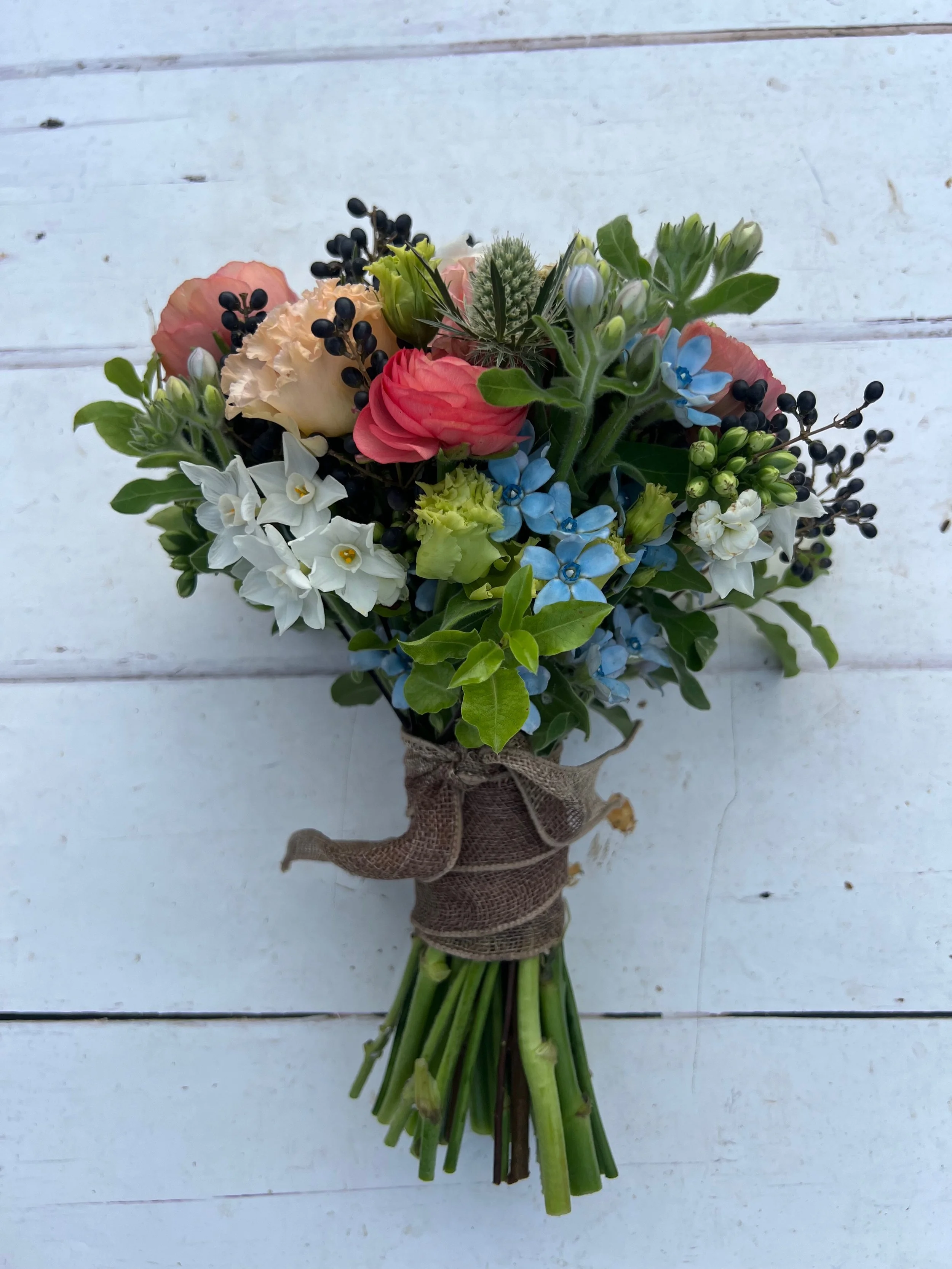 Bouquet of mixed flowers including white, pink, and blue blooms, with green foliage, tied with burlap ribbon, on a white wooden surface.