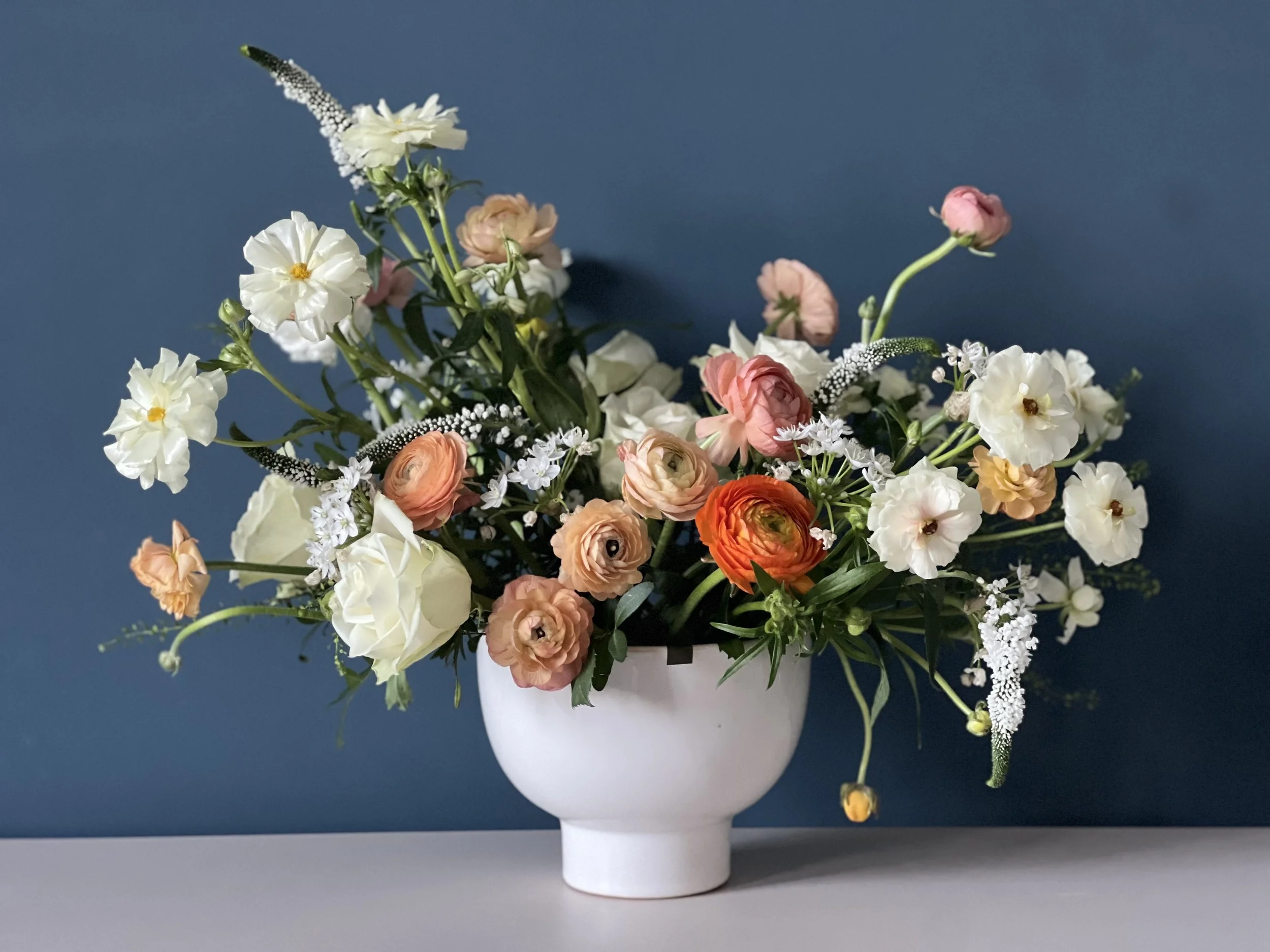 A white vase with a bouquet of mixed flowers in white, peach, pink, and orange, placed on a surface with a dark blue background.