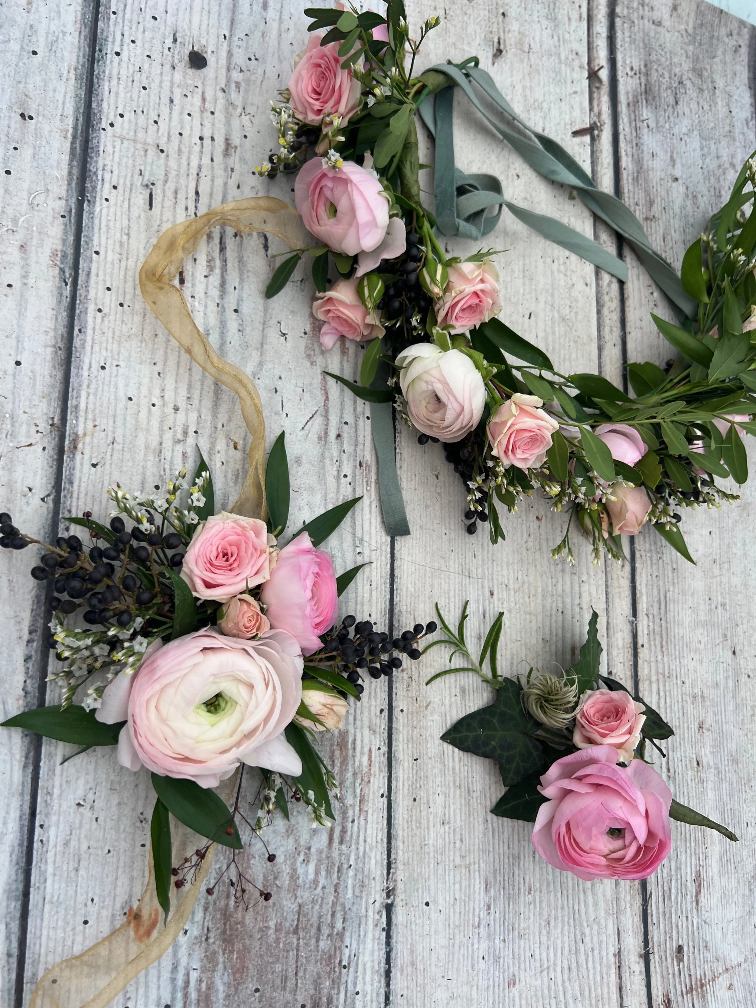 A floral crown and a boutonniere made with pink roses, white ranunculus, greenery, black berries, and ribbons laid on a white wooden surface.