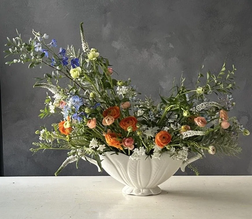 Elegant floral arrangement in a white ceramic bowl with colorful flowers including orange roses, pink ranunculus, blue delphiniums, and white accents, set against a dark gray background.