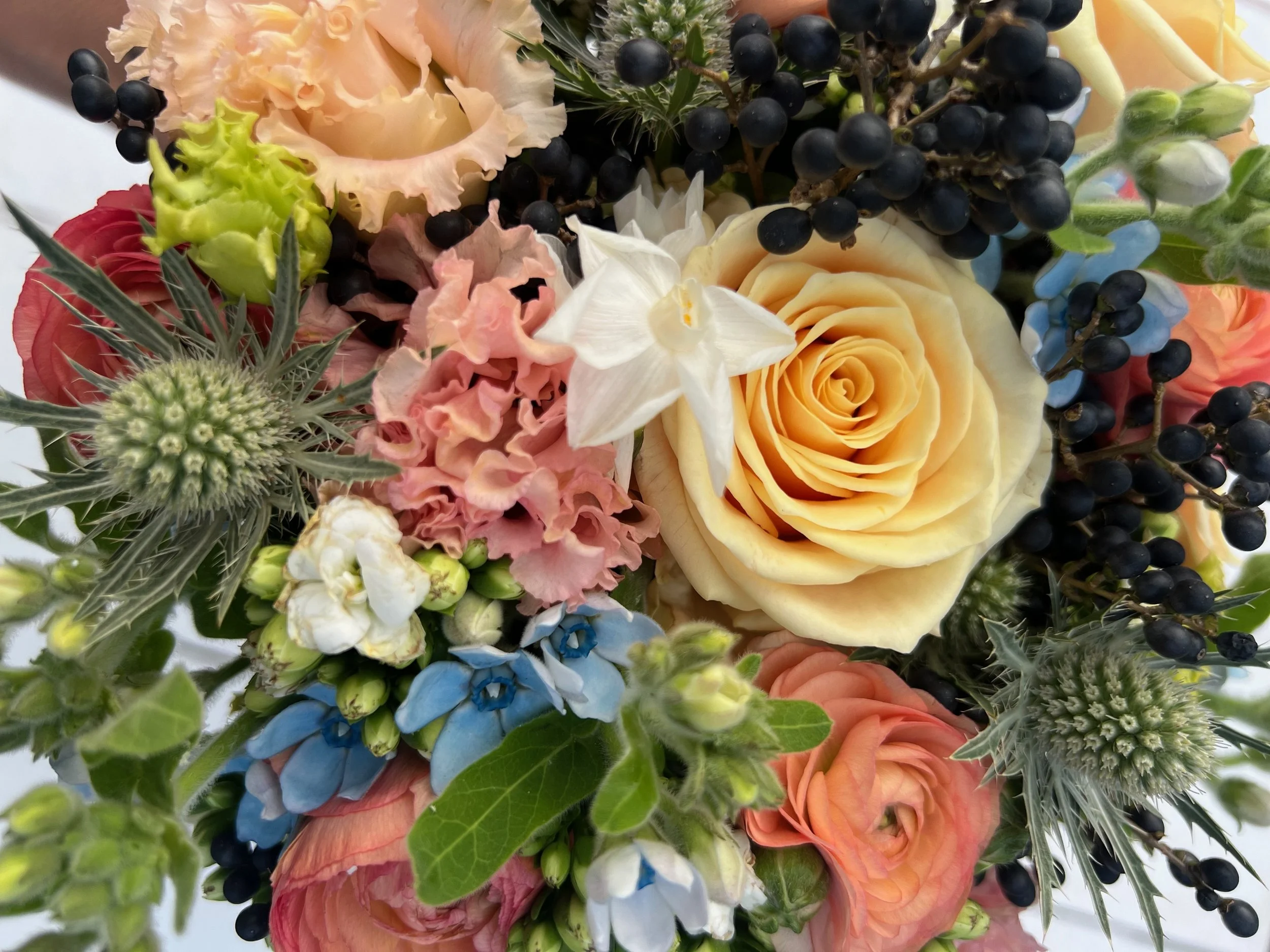 A colorful bouquet of flowers including yellow roses, pink roses, white orchids, blue delphiniums, and various green foliage and berries.