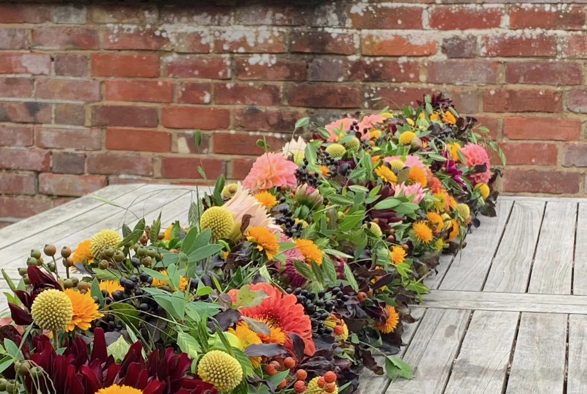 A long floral arrangement with various colorful flowers, leaves, and berries, placed on a weathered wooden table against a brick wall.